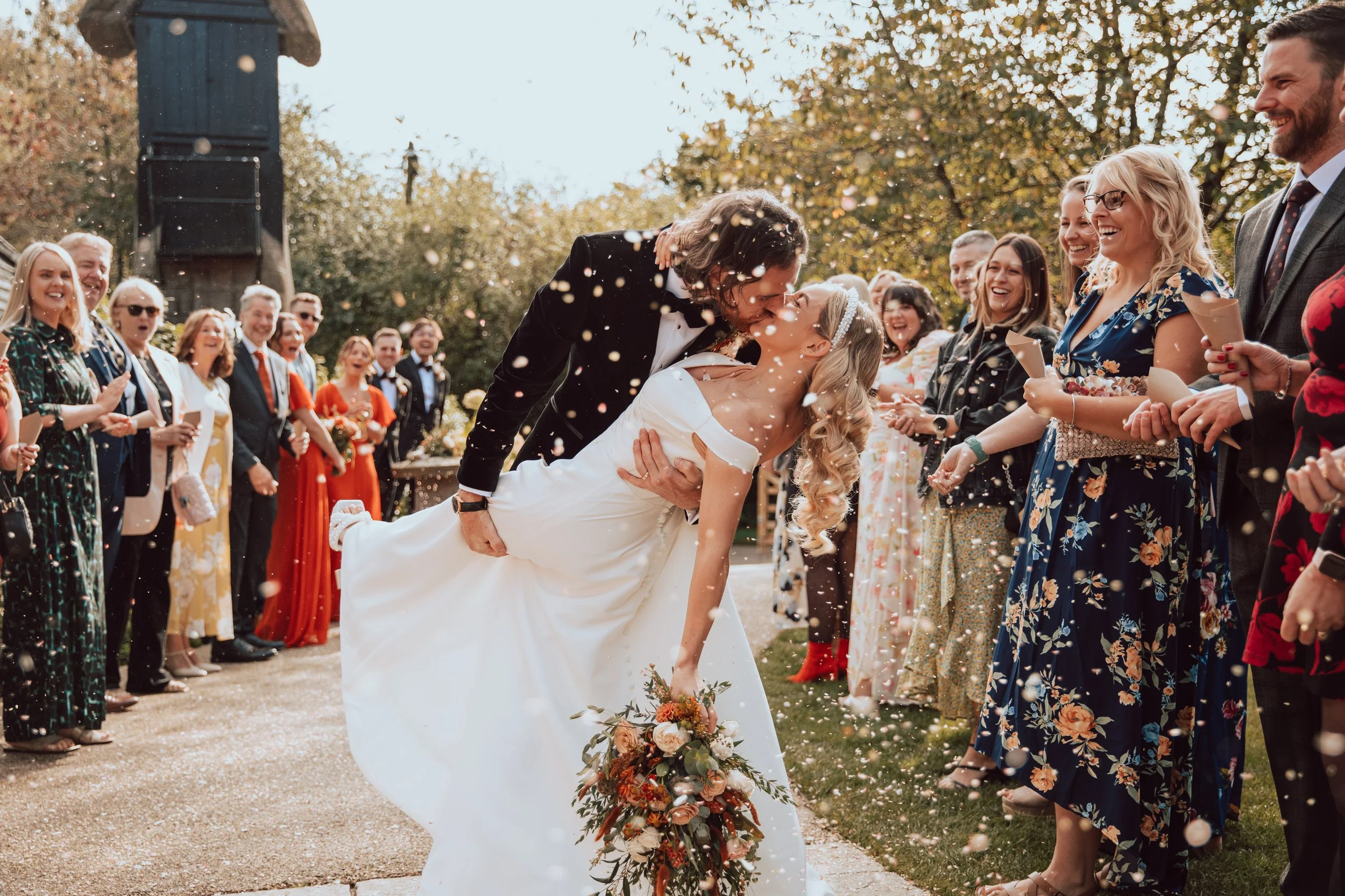 A newlywed couple kissing during an outdoor wedding celebration, surrounded by friends and family, with confetti falling and trees in the background.