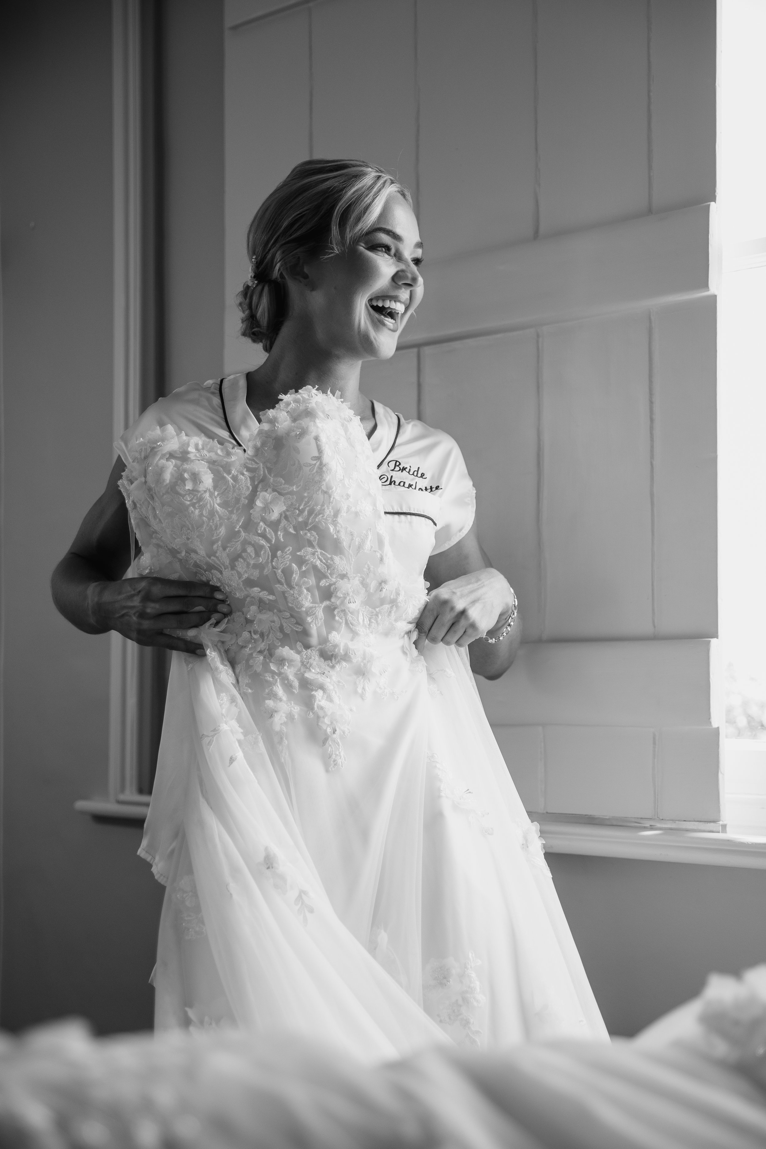 A woman in a wedding dress joyful and smiling, standing by a window with natural light, in a room with wooden paneled walls.