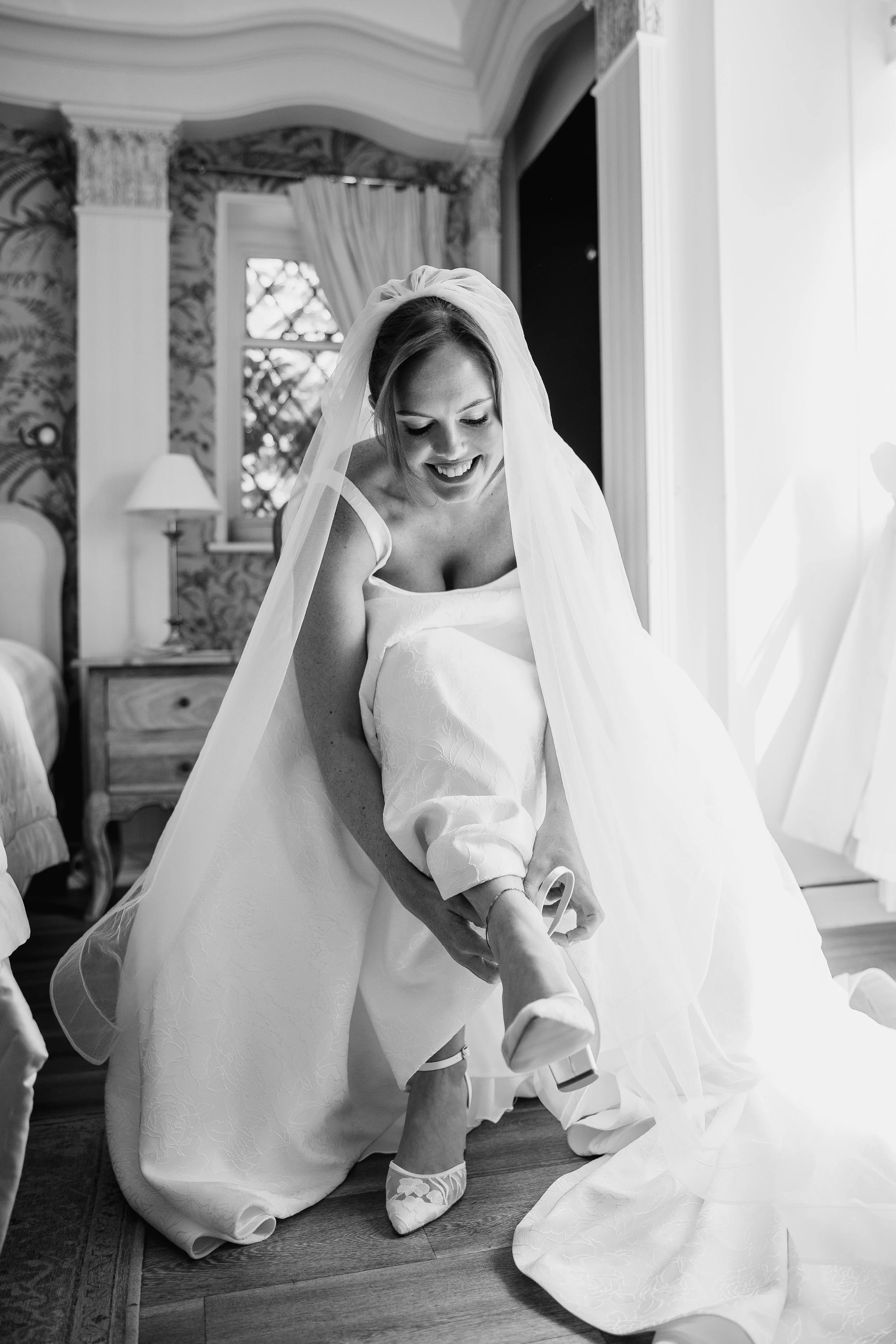 A woman in a wedding dress smiling, as she puts on her shoe, in a room with floral wallpaper, a window with curtains, and a bedside lamp.