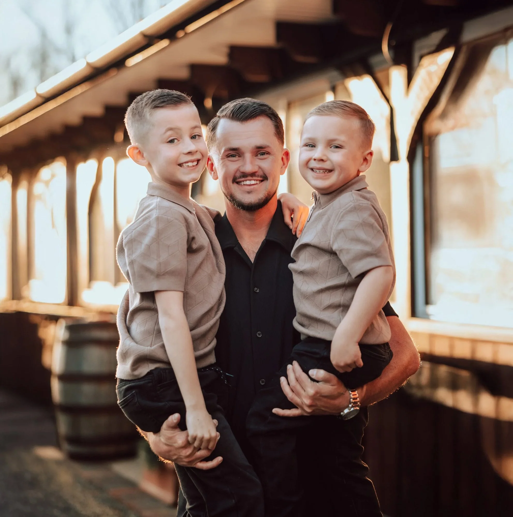 A man is holding two children, one in each arm, standing outdoors near a wooden structure during sunset. The children are smiling and the man is smiling, with warm light illuminating their faces. Suffolk Wedding Photographer / Essex Wedding Photos