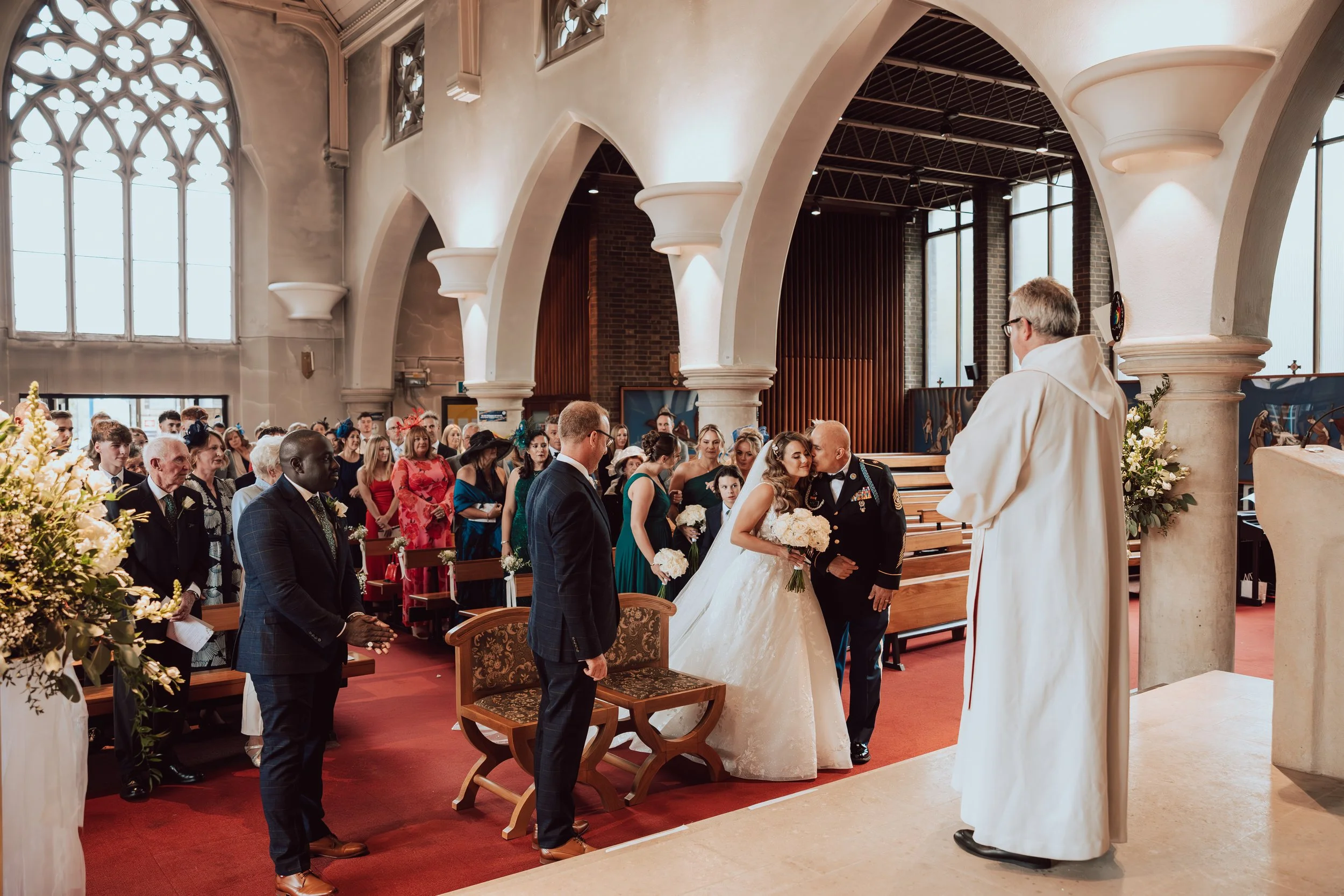 A wedding ceremony inside a church with a bride and groom standing in front of a priest. Guests are seated in pews, and the church has tall, arched windows and ornate columns.