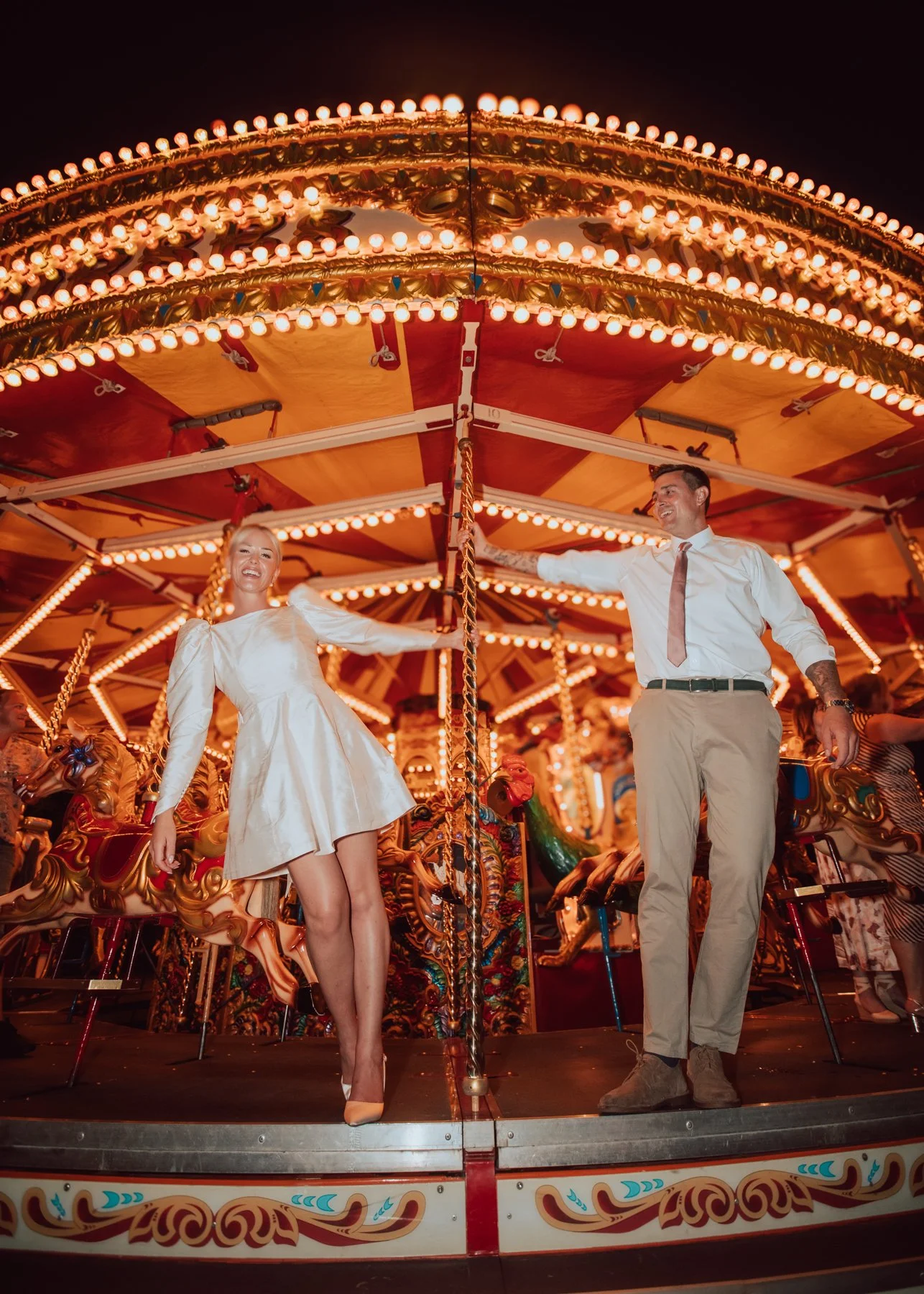 A woman in a white dress and a man in a white shirt and beige pants standing on a carousel at night, with happy expressions.