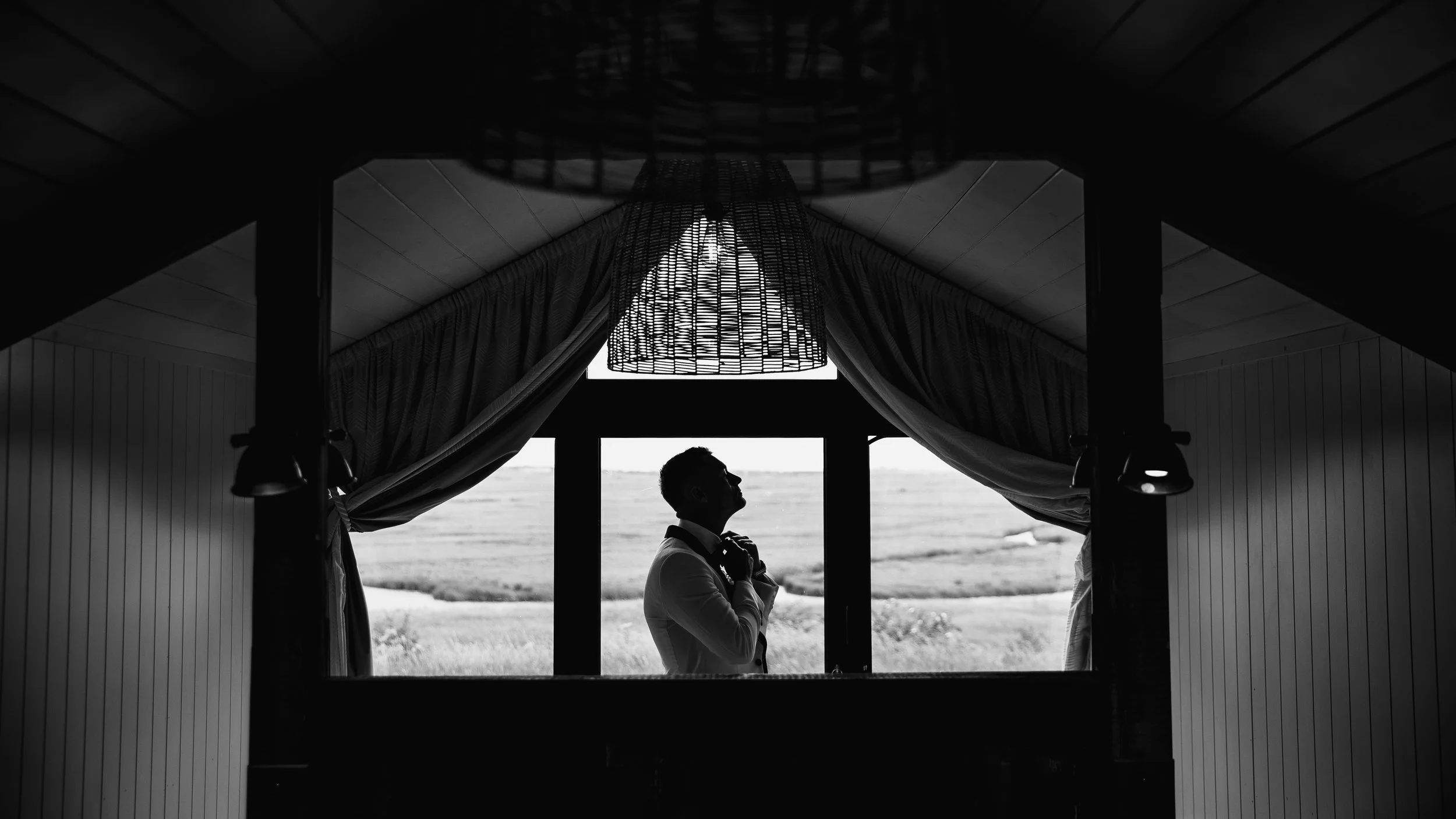Silhouette of a man adjusting his tie while looking out a window framed by curtains, with a landscape visible outside.