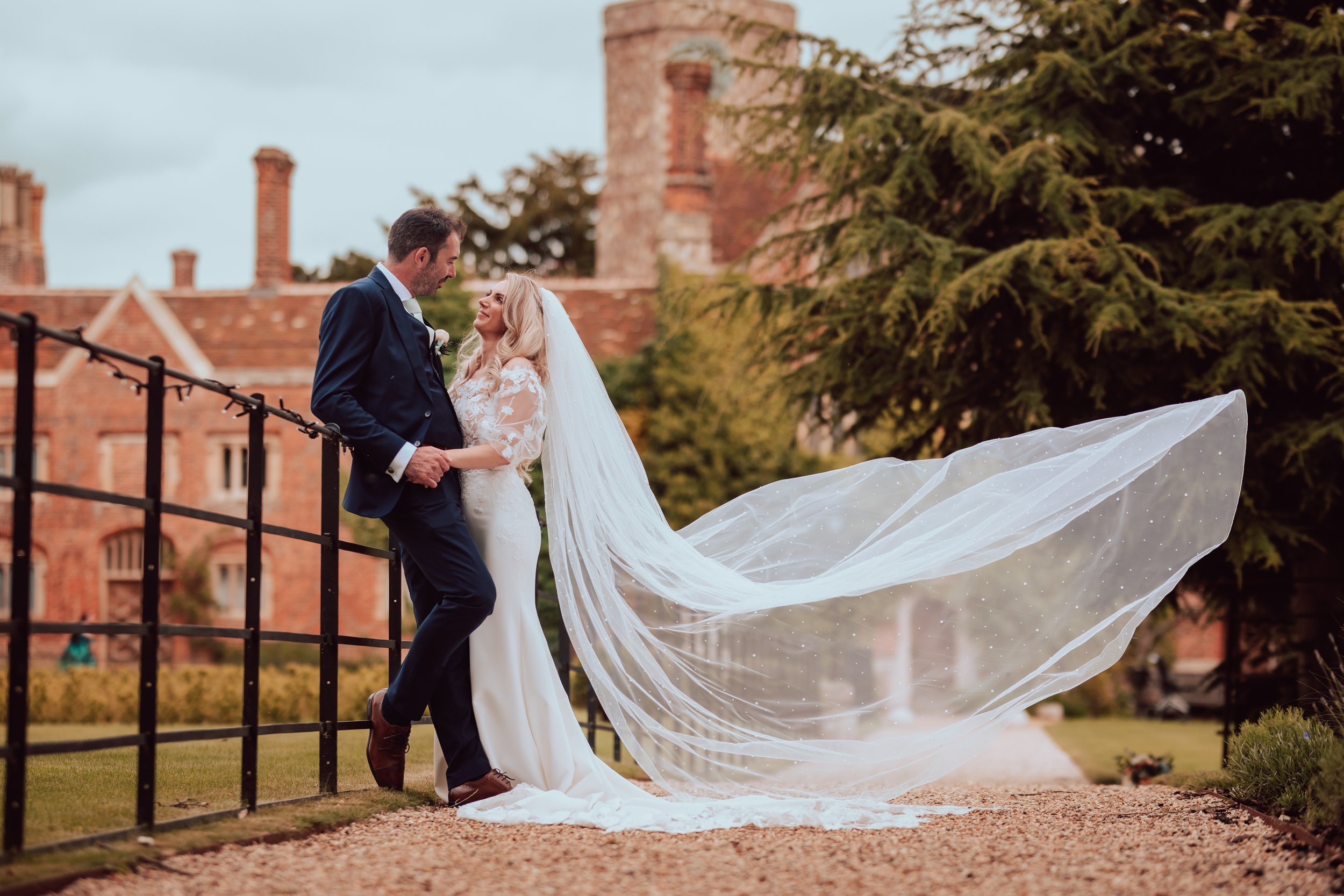 A bride and groom holding hands and looking at each other outside during a wedding, with the groom in a dark suit and the bride in a white wedding dress with a train and veil, near a black fence and a brick building with chimneys in the background.
