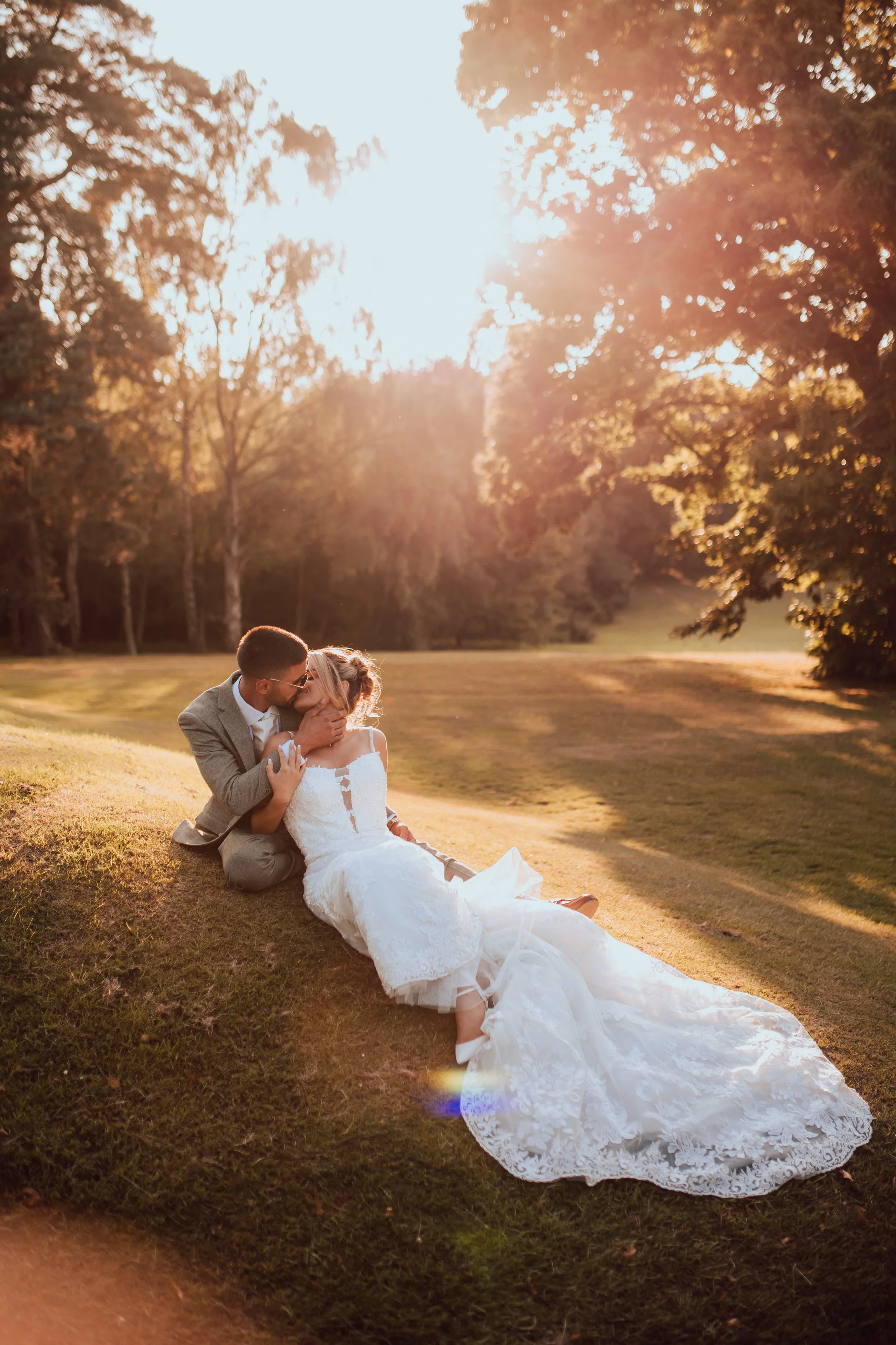 A couple sitting on the grass during sunset in a park, with the man kissing the woman's forehead and the woman in a white wedding dress and the man in a grey suit.