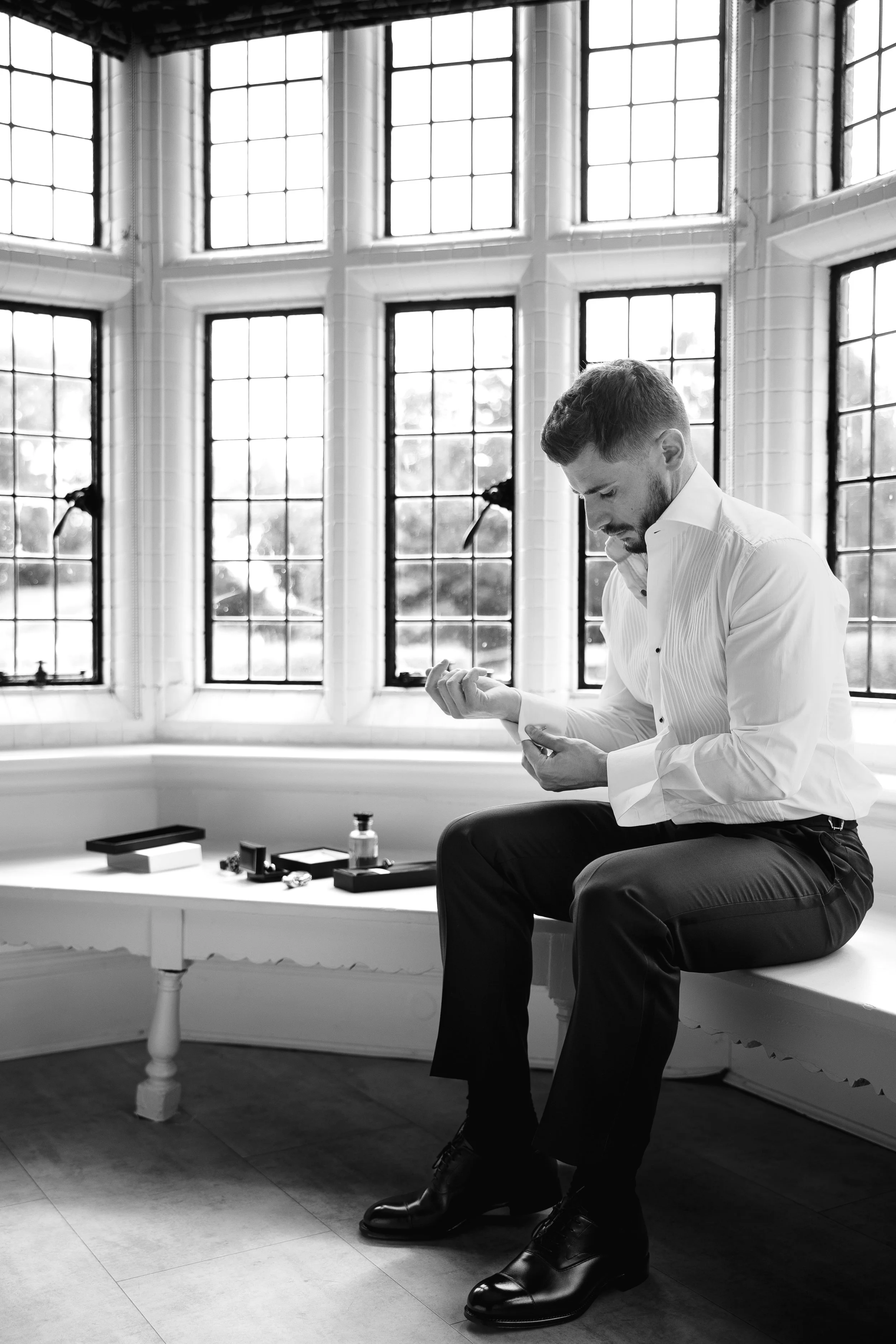 A man in a white dress shirt and dress shoes is sitting on a windowsill, looking down and adjusting his cufflinks. The room features large, multi-pane windows with bright sunlight streaming in. A table nearby holds a few small items, including a bottle and some boxes.