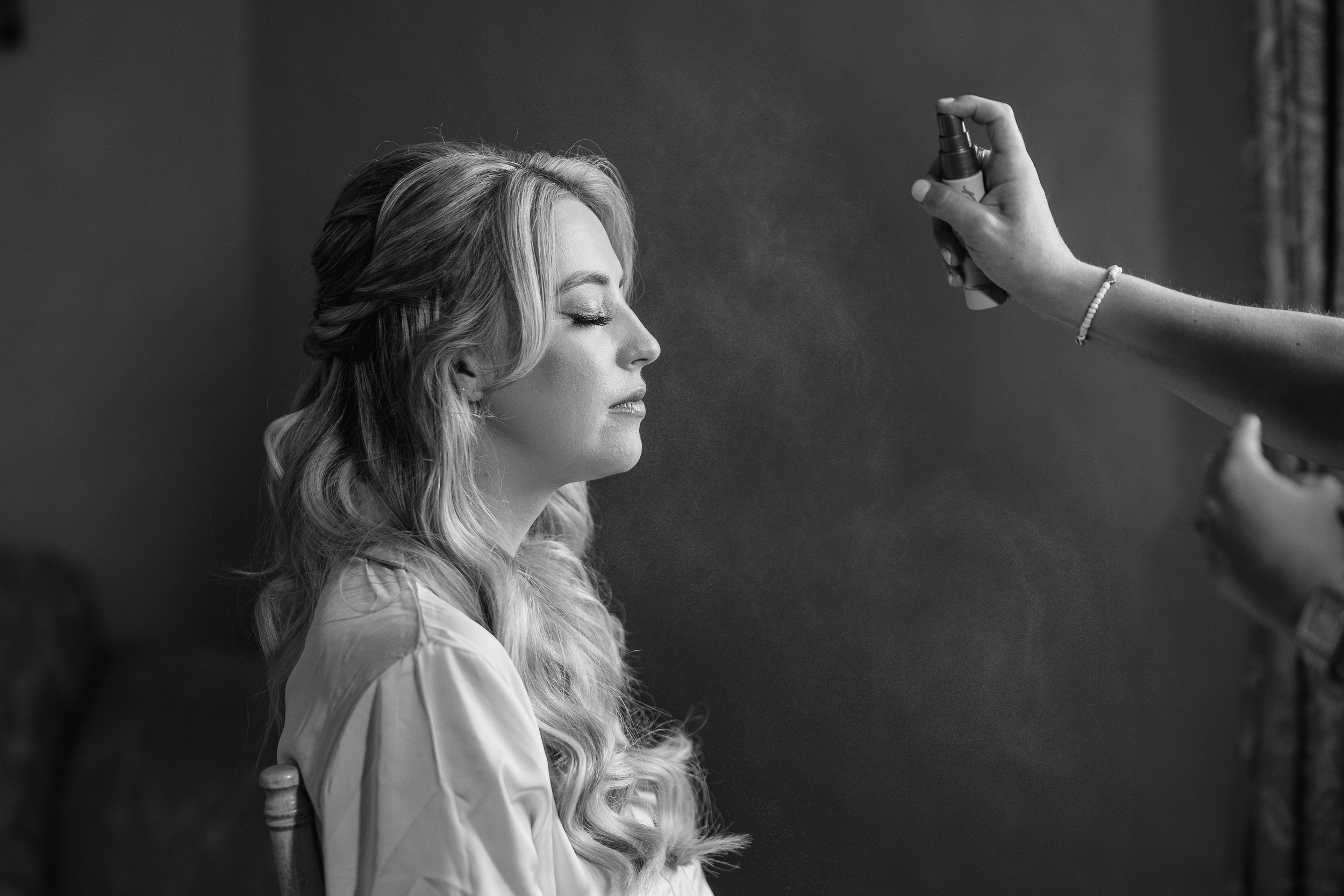 Black and white photo of a woman with long, wavy hair sitting with her eyes closed as a makeup artist applies spray to her face.