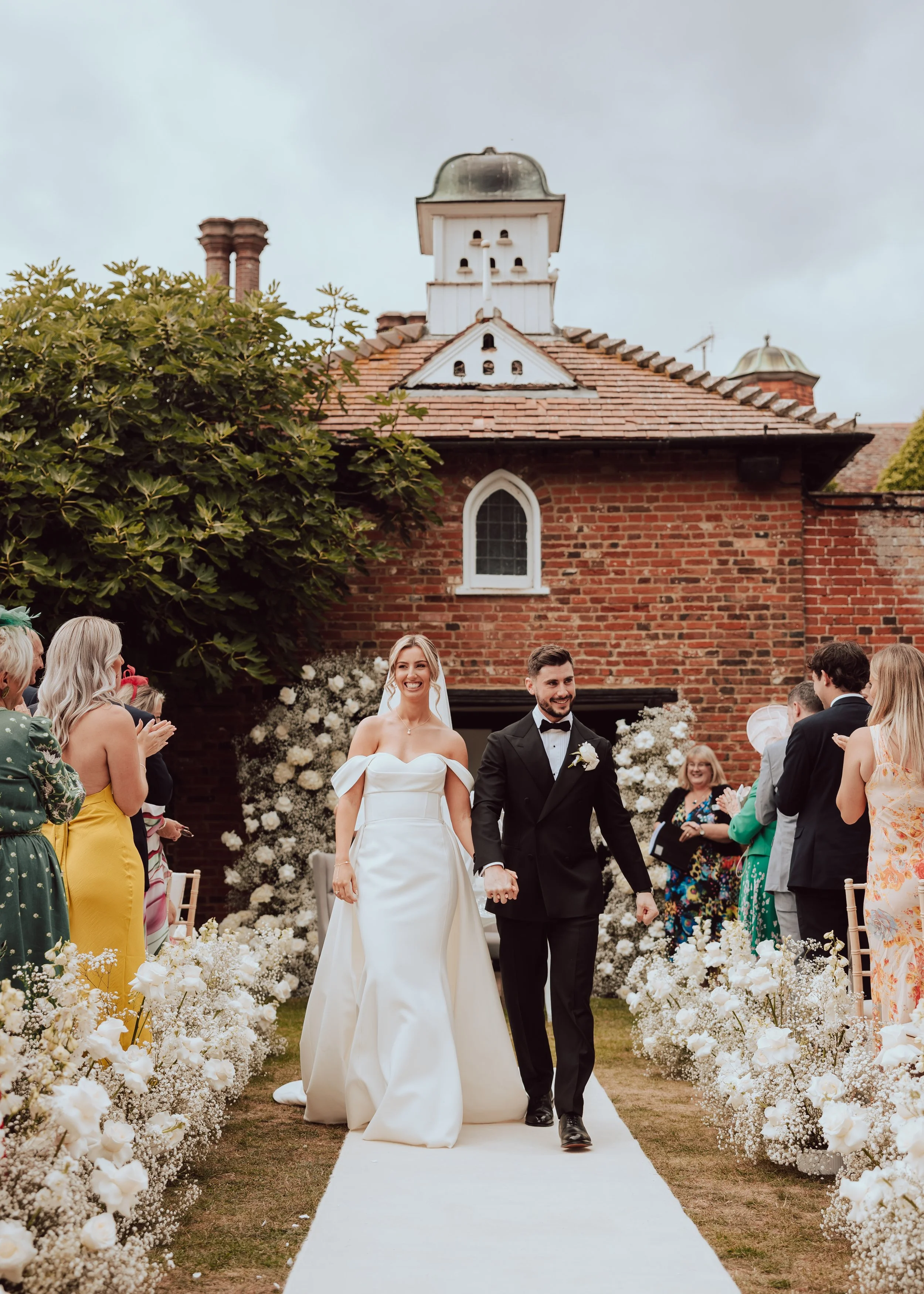 A newly married couple walking down an outdoor aisle lined with white flowers at their wedding ceremony, with guests on both sides, in front of a red brick building with a small tower and chimneys.