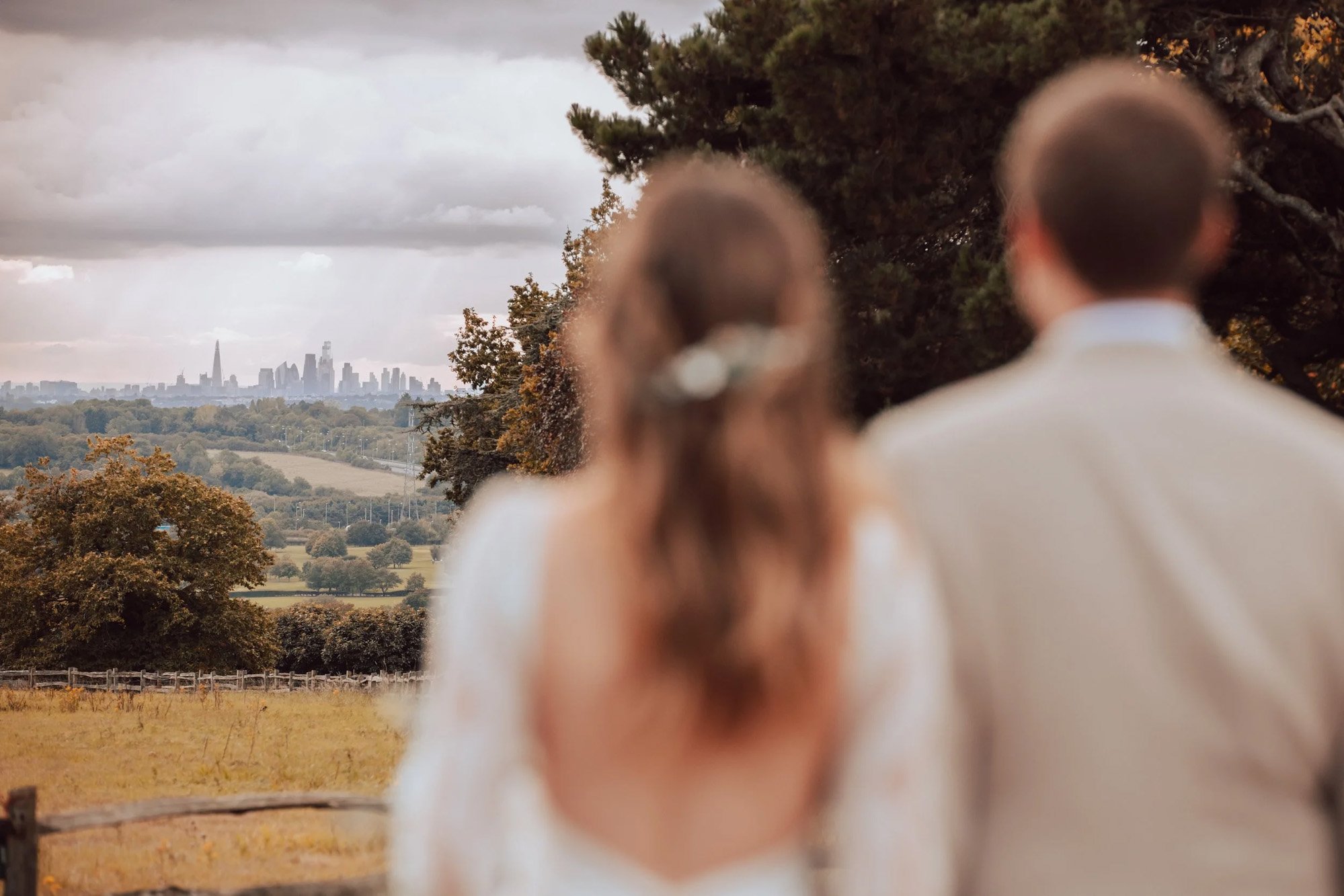 Blurred photo of two people, a woman and a man, standing outdoors with a city skyline in the background. The woman has brown hair tied back and is wearing earrings; the man has short hair and is wearing a white shirt. Essex Wedding Photographer