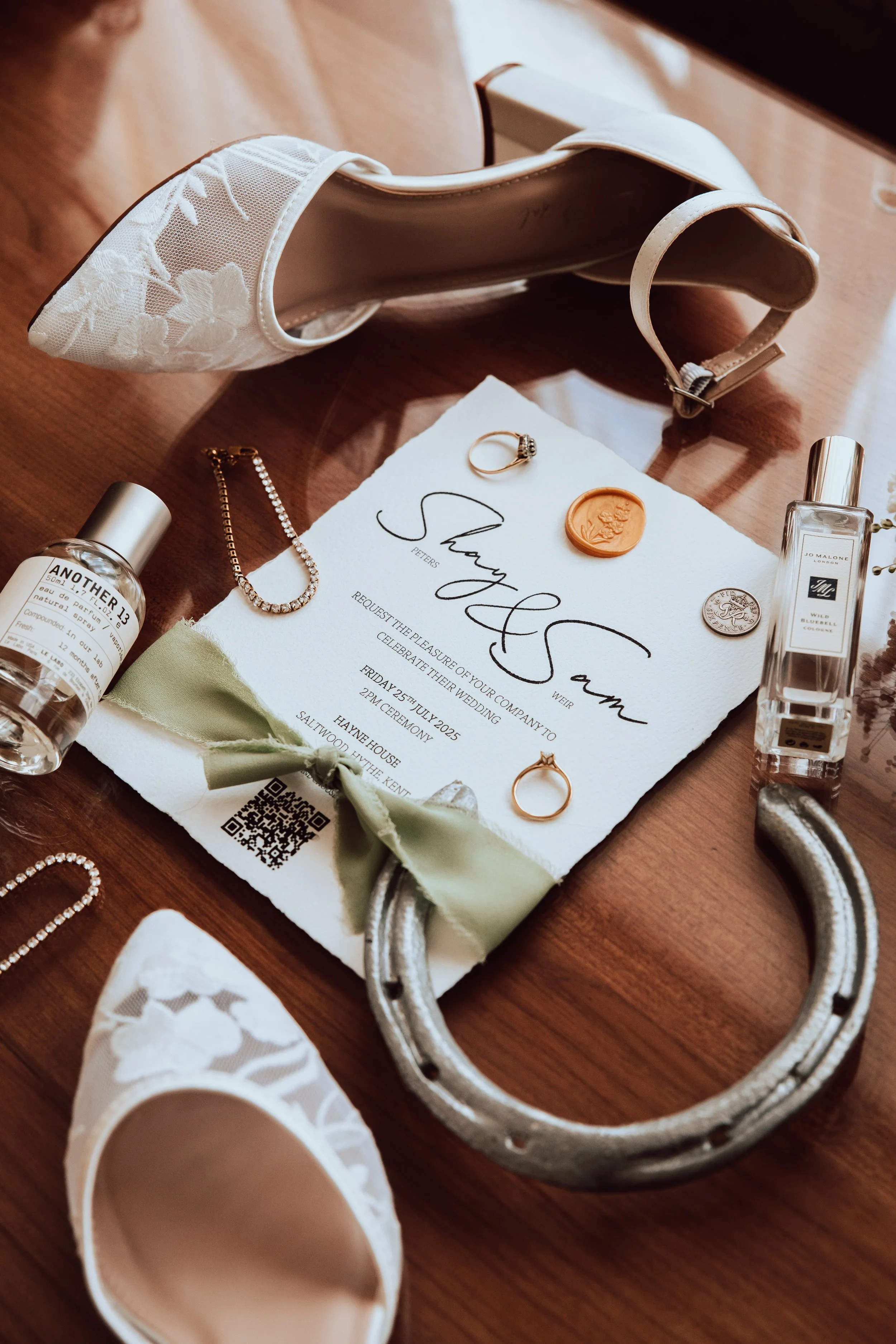 A pair of white embroidered high-heeled shoes, a wedding invitation, jewelry including rings and a necklace, hand sanitizer bottles, and a white lace garter arranged on a wooden surface.