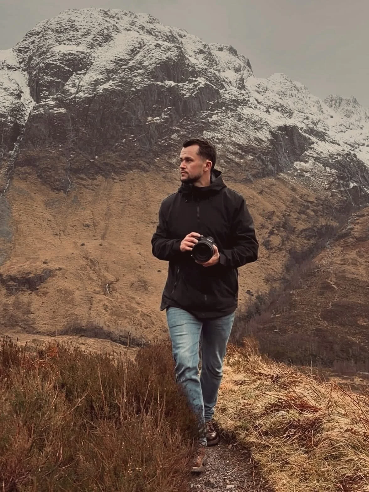 A man holding a camera stands outdoors in front of a mountainous landscape with snow-capped peaks and brownish terrain. Photographer Liam Gillan in Glencoe Scotland