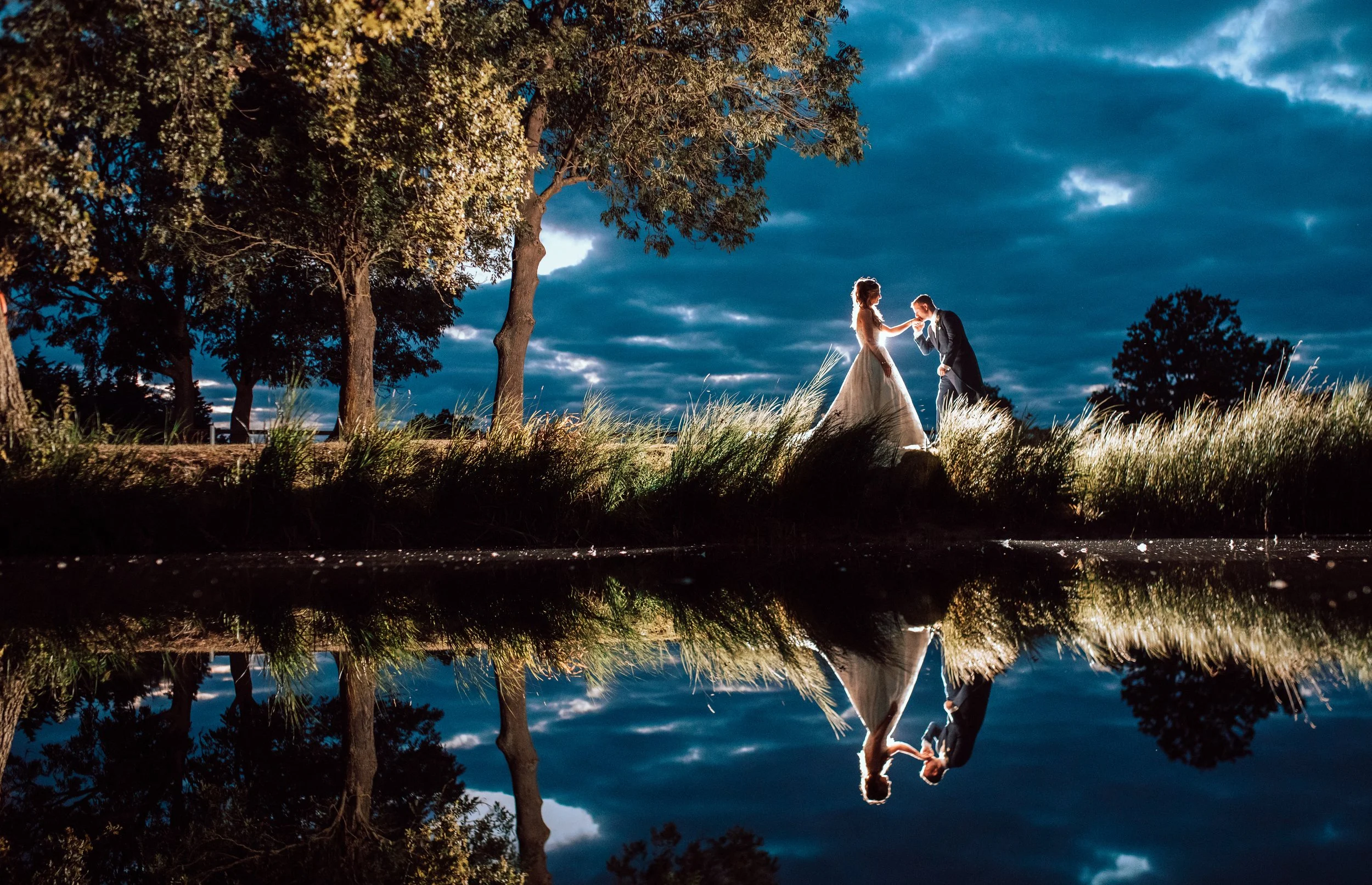 A bride and groom at sunset, standing on grass near a tree, with their reflection visible in a body of water, under a partly cloudy sky.
