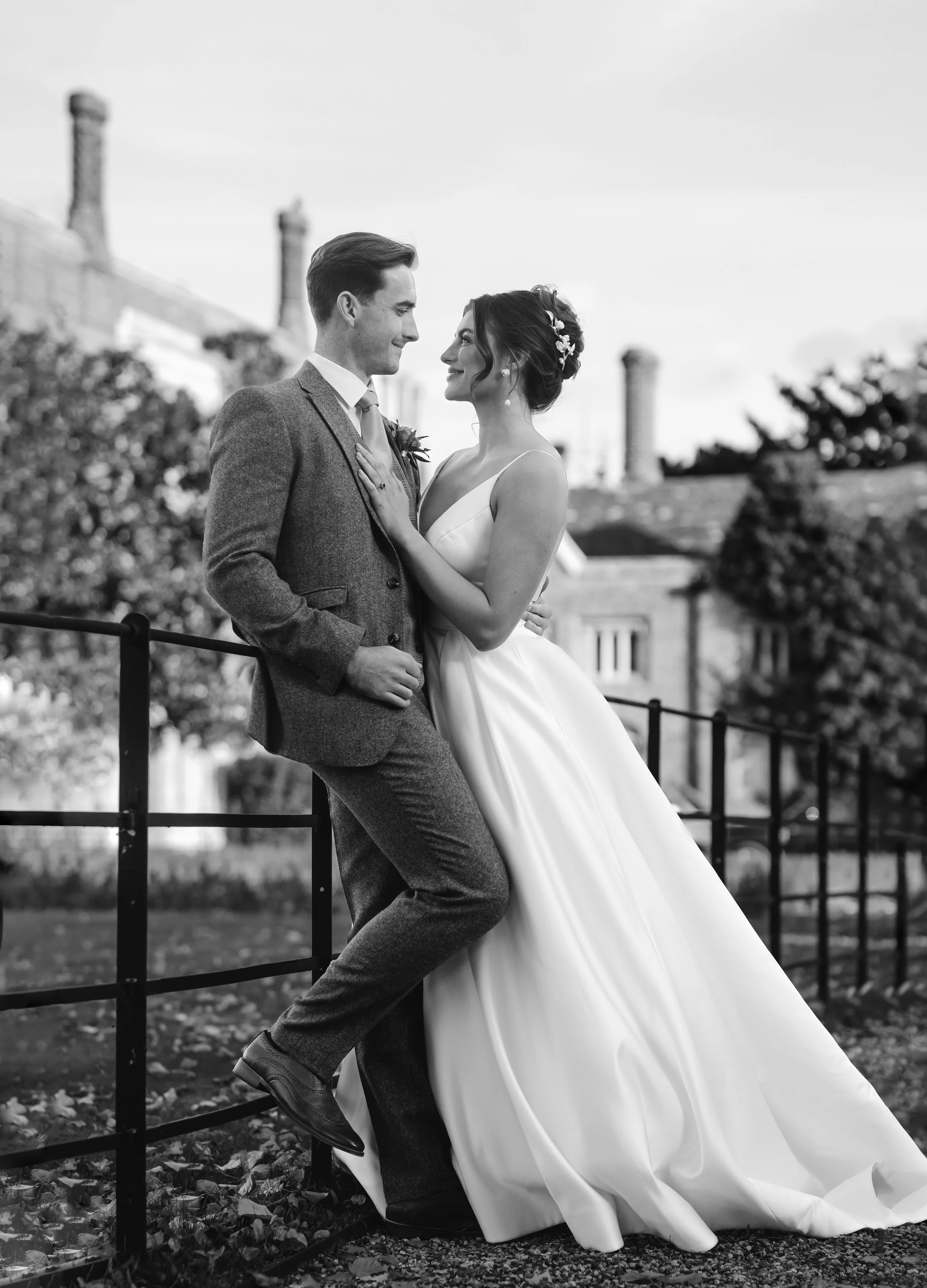 A black and white photo of a bride and groom on their wedding day. The groom is wearing a suit and tie, and the bride is in a wedding dress with floral hair accessories. They are standing outdoors near a railing with buildings and trees in the background, gazing into each other's eyes.