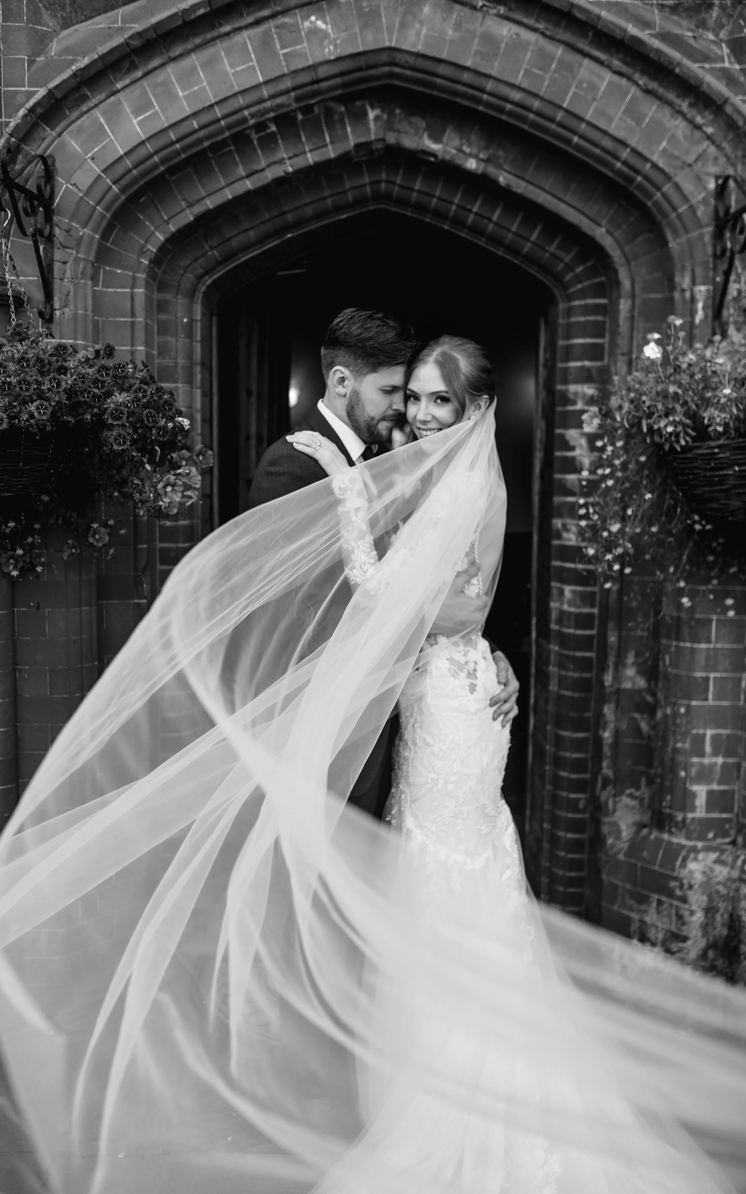 A bride and groom dressed in wedding attire posing under a brick archway, with the bride's veil flowing in the wind.
