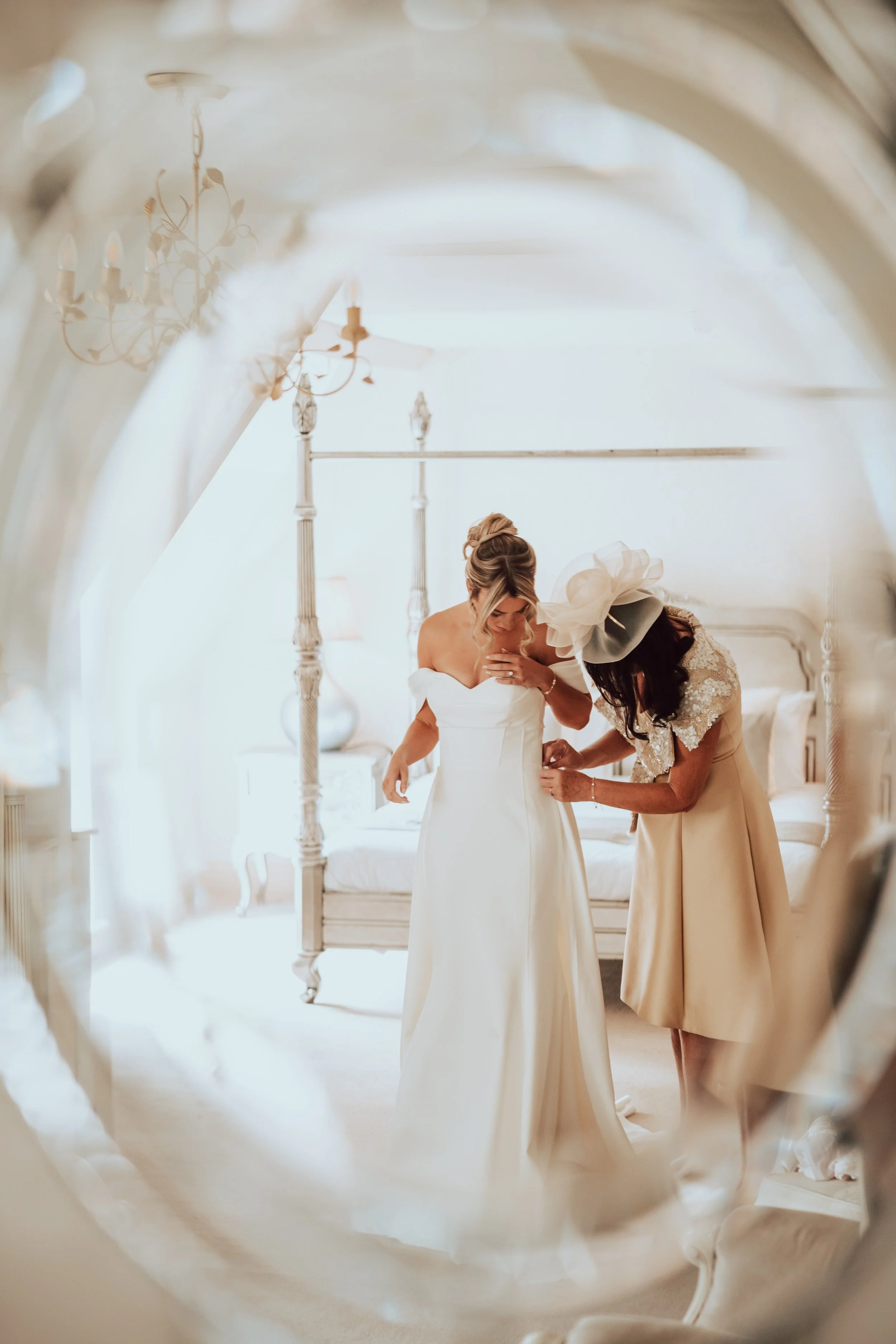 A bride in a white wedding dress getting ready, assisted by another woman in a beige dress with a large white hat, in a bright, elegant room with chandeliers and a four-poster bed.