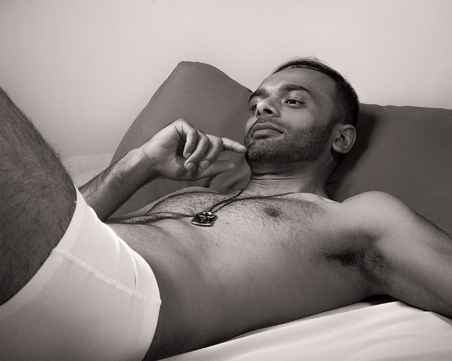 Black and white photo of a man lying down, wearing underwear and a necklace, posed in a relaxed manner.