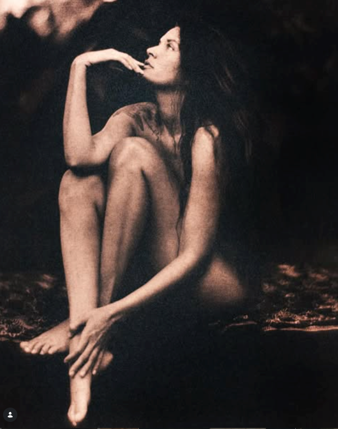 A woman sitting nude with her legs crossed, resting her head on her hand, with long dark hair flowing around her shoulders, in a dark, moody setting.