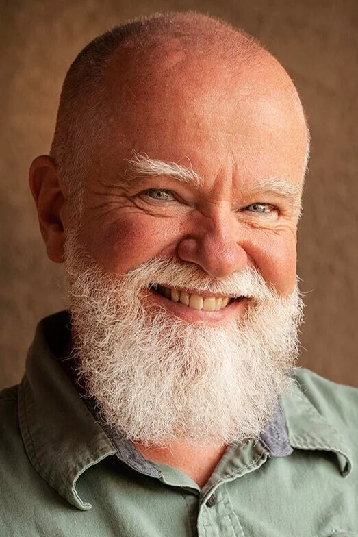 Close-up of a smiling elderly man with a white beard and white eyebrows, wearing a green shirt, against a plain brown background.