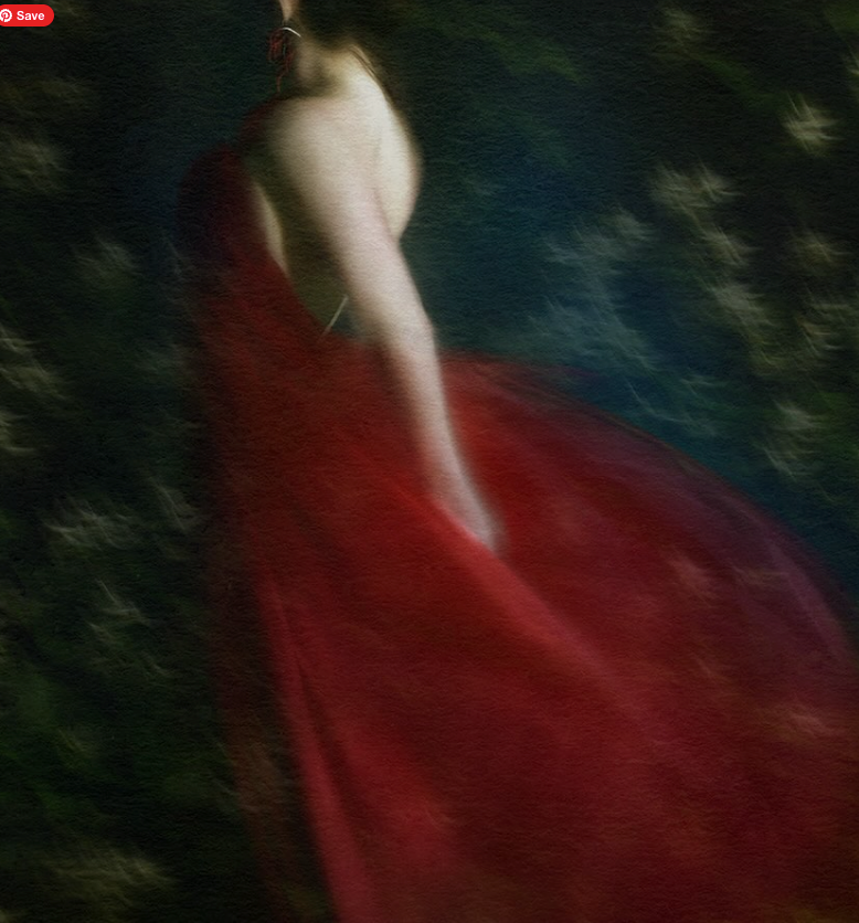 A woman with red hair wearing a flowing red dress, standing outdoors in a blurry, forested background.