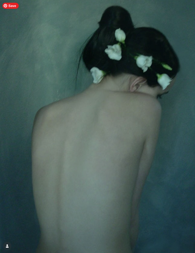A woman with dark hair decorated with white flowers, sitting with her back to the camera and her head tilted down, revealing her bare back and shoulders against a muted background.