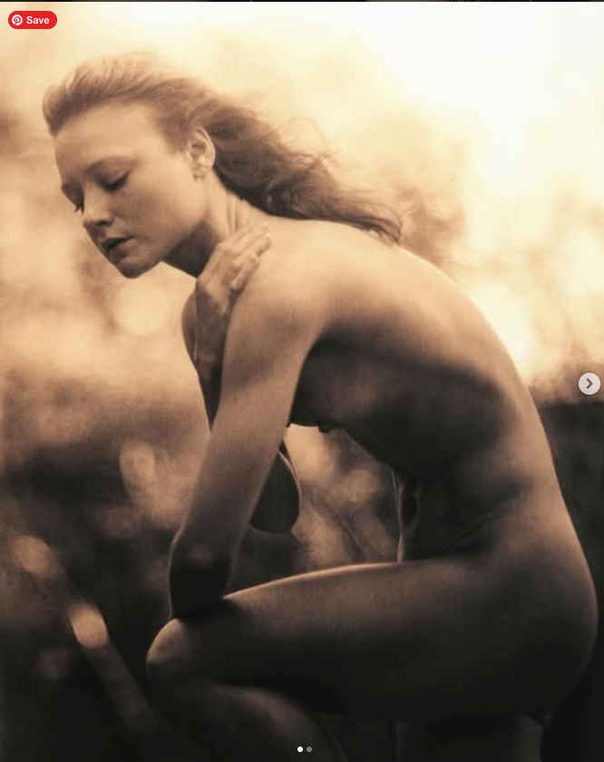 A black-and-white photograph of a nude woman with long flowing hair, sitting outdoors with her eyes closed and head slightly bowed, in a contemplative pose.