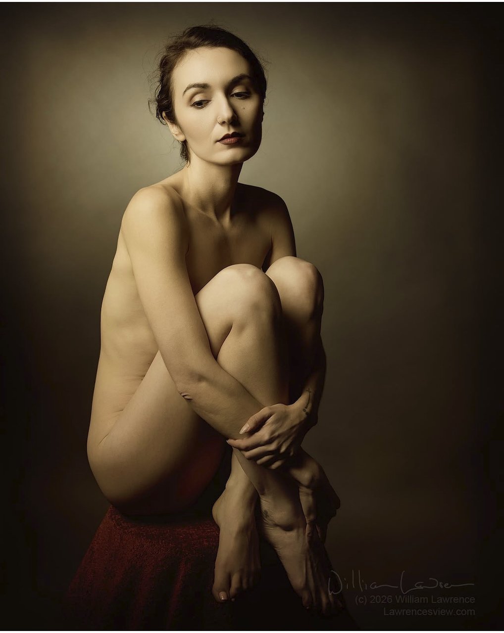 A nude woman with dark brown hair seated on a red cloth-covered surface against a dark background, with her arms wrapped around her knees, looking down and to the side with a contemplative expression.