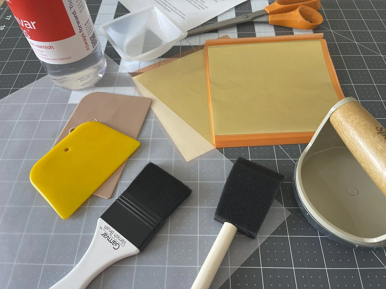 Crafting supplies including a bottle of clear varnish, a white plastic spoon, orange scissors, a square orange picture frame with yellow paper inside, yellow and pink paint swatches, a black paintbrush, black sponge brush, a round tape roller, and a black foam brush on a cutting mat.