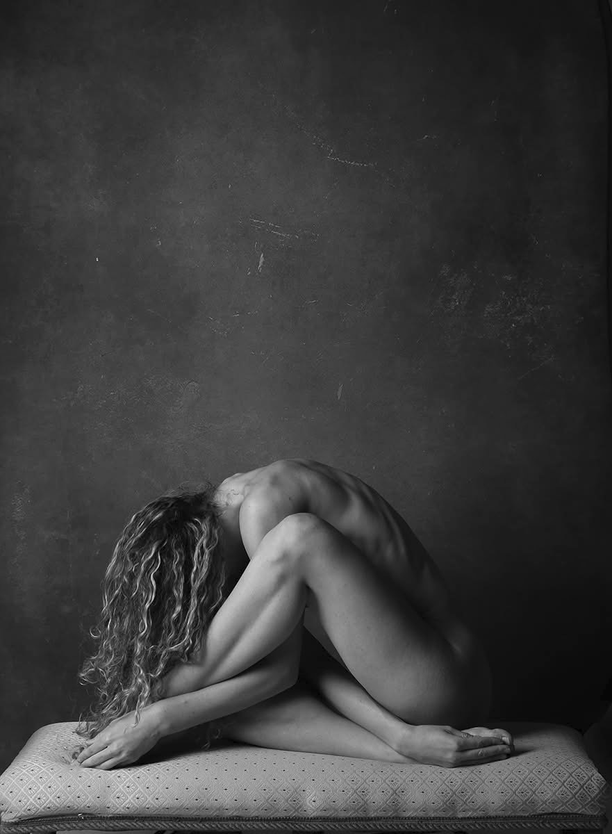 Black and white photo of two nude women intertwined on a padded surface against a textured dark wall, with one woman with curly hair and the other with straight hair.