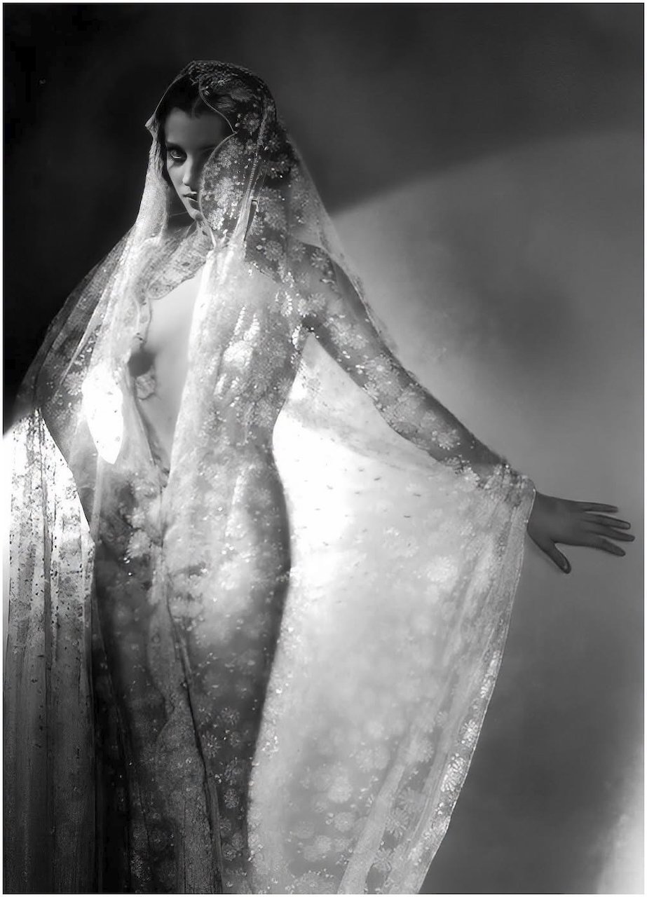 Black and white photo of a woman with dark hair, partially covered by a sheer, patterned fabric draped over her head and shoulders, posing with her left hand extended outward.