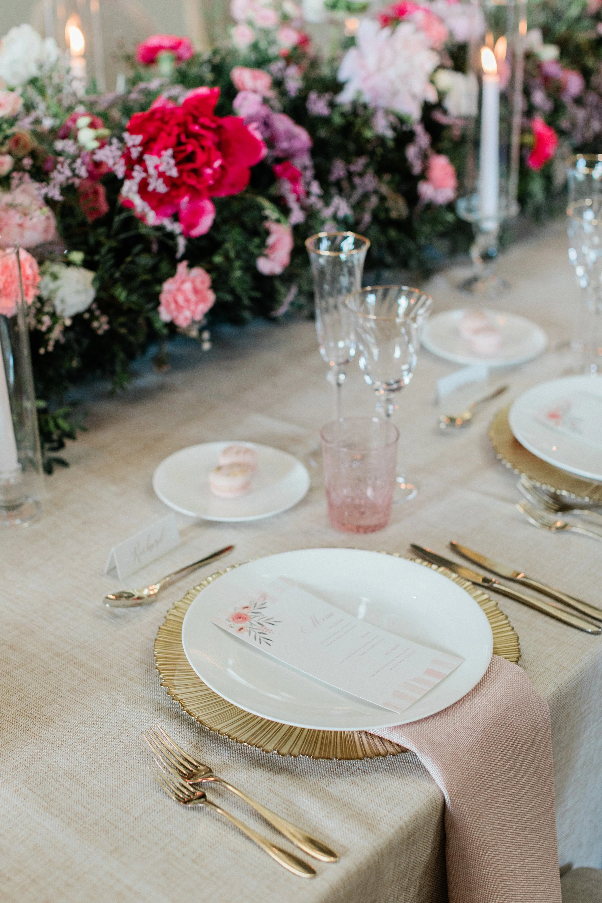 Elegant wedding table setting with gold-rimmed plates, pink napkin, floral menu card, glassware, and a floral centerpiece with pink, white, and red flowers and candles.
