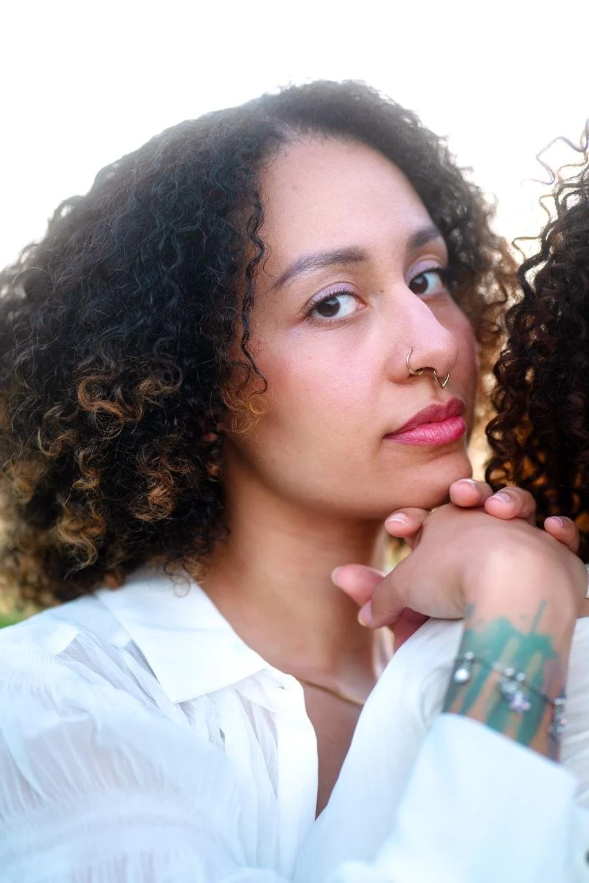 Close-up of Michelliink Michelli Nyasha Marimo Kristensen with curly hair and a nose ring, resting her chin on her hand, wearing a white shirt. owner of MIIM