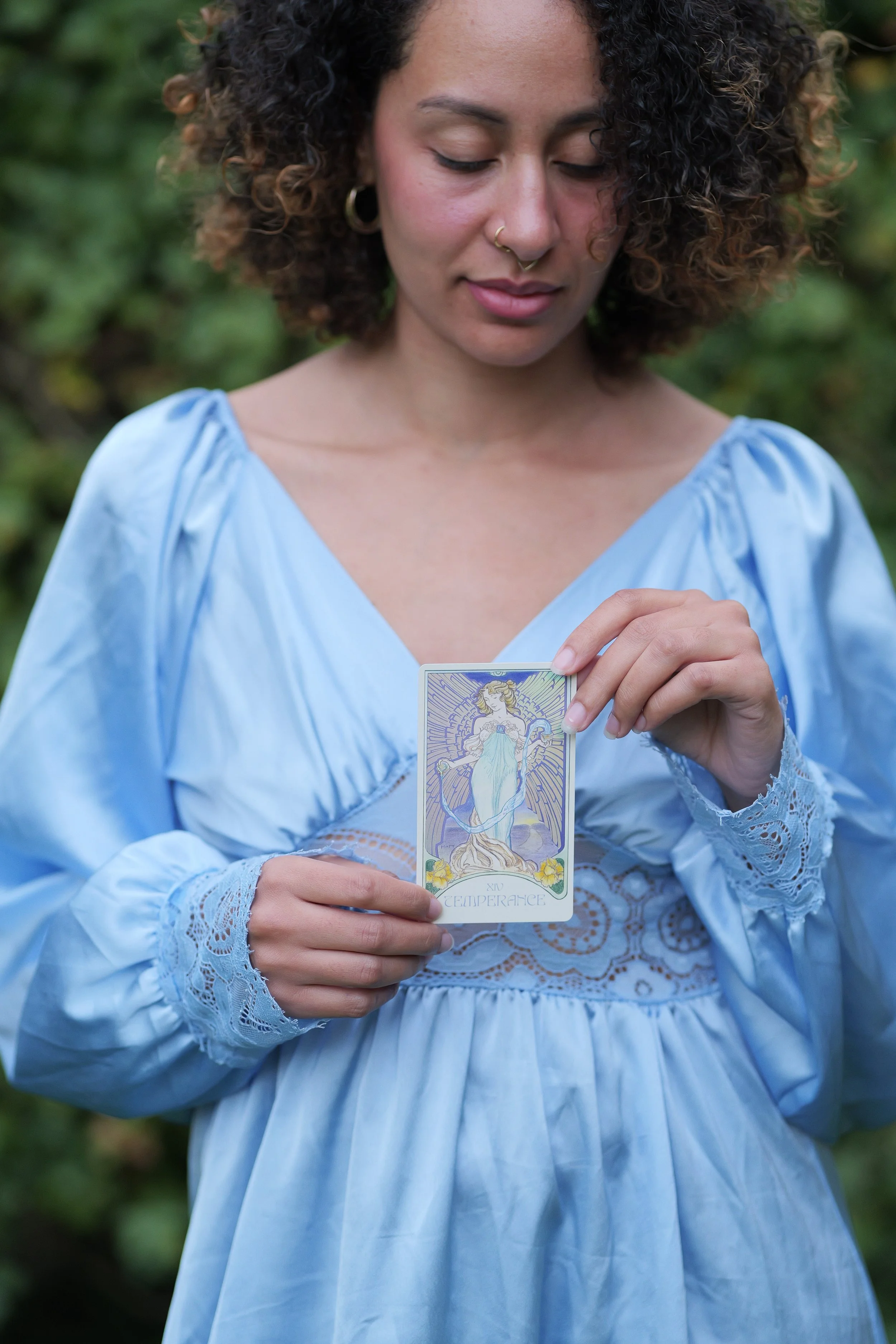 Michelliink Michelli Nyasha Marimo Kristensen in a light blue dress with lace details is holding a Tarot card titled 'Temperance', standing outdoors with greenery in the background. owner of MIIM