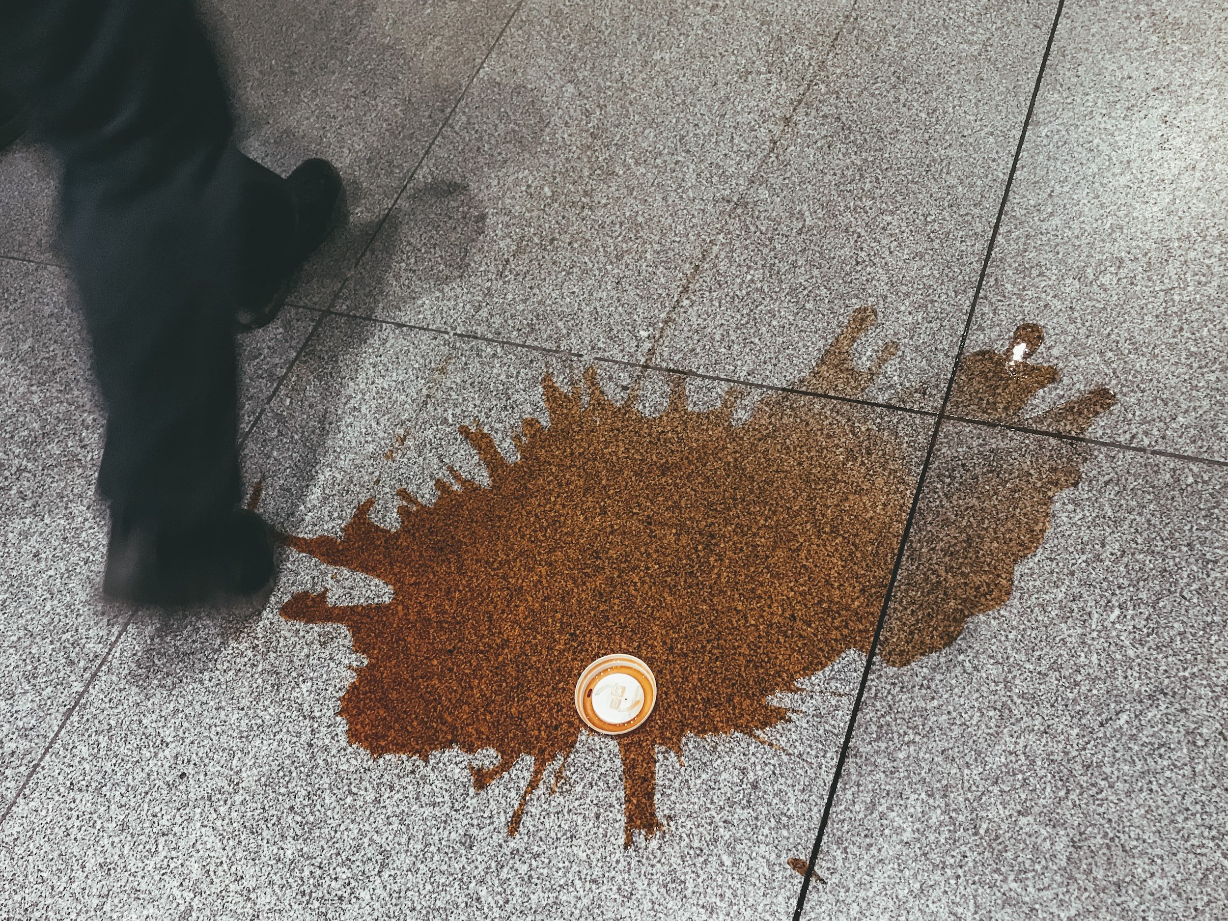 a person walking by a puddle of spilled coffee with a plastic cup lid