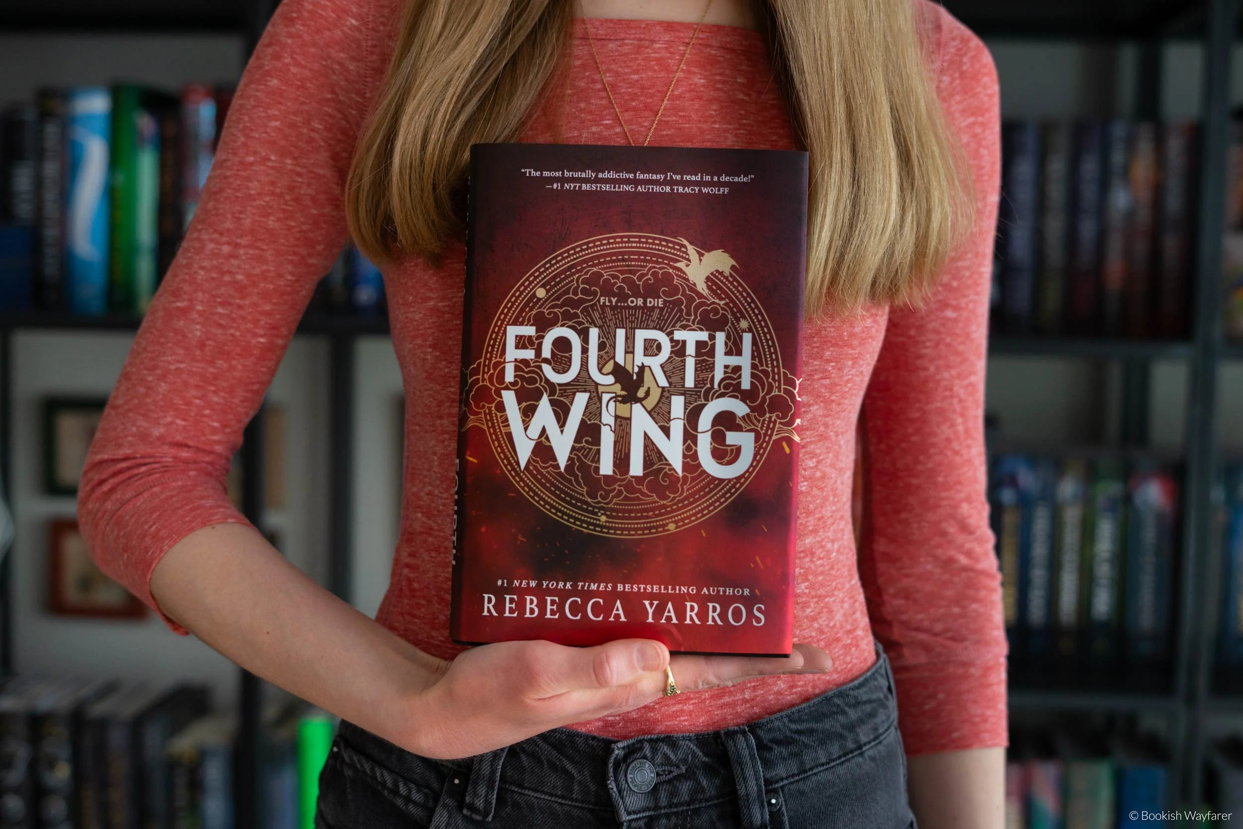 A woman in an orange shirt holds the book Fourth Wing in front of her