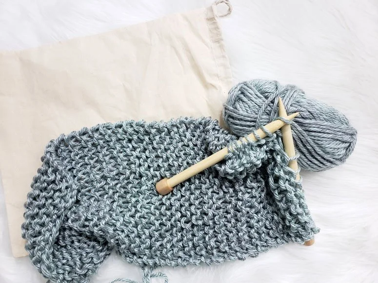 A ball of yarn and knitting needles with a partially completed scarf laid flat atop a white background