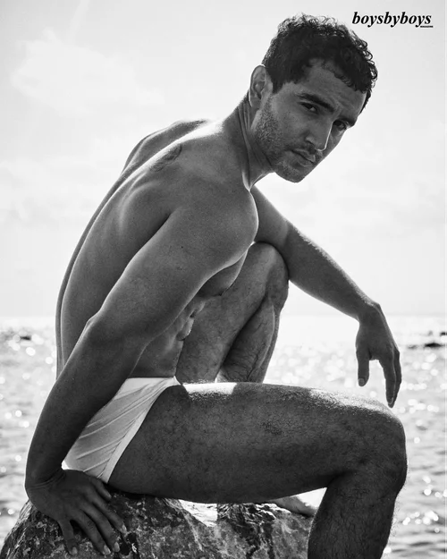 Black and white photo of a man in white swimwear sitting on a rock by the sea.