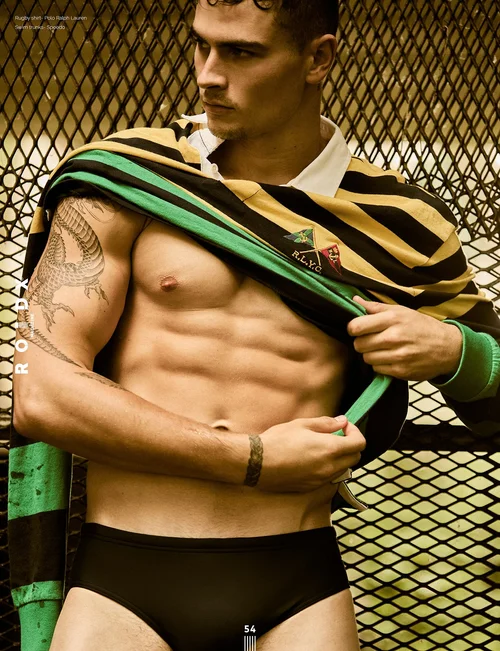 Shirtless male model wearing striped rugby shirt and black swim trunks, standing in front of metal fence.
