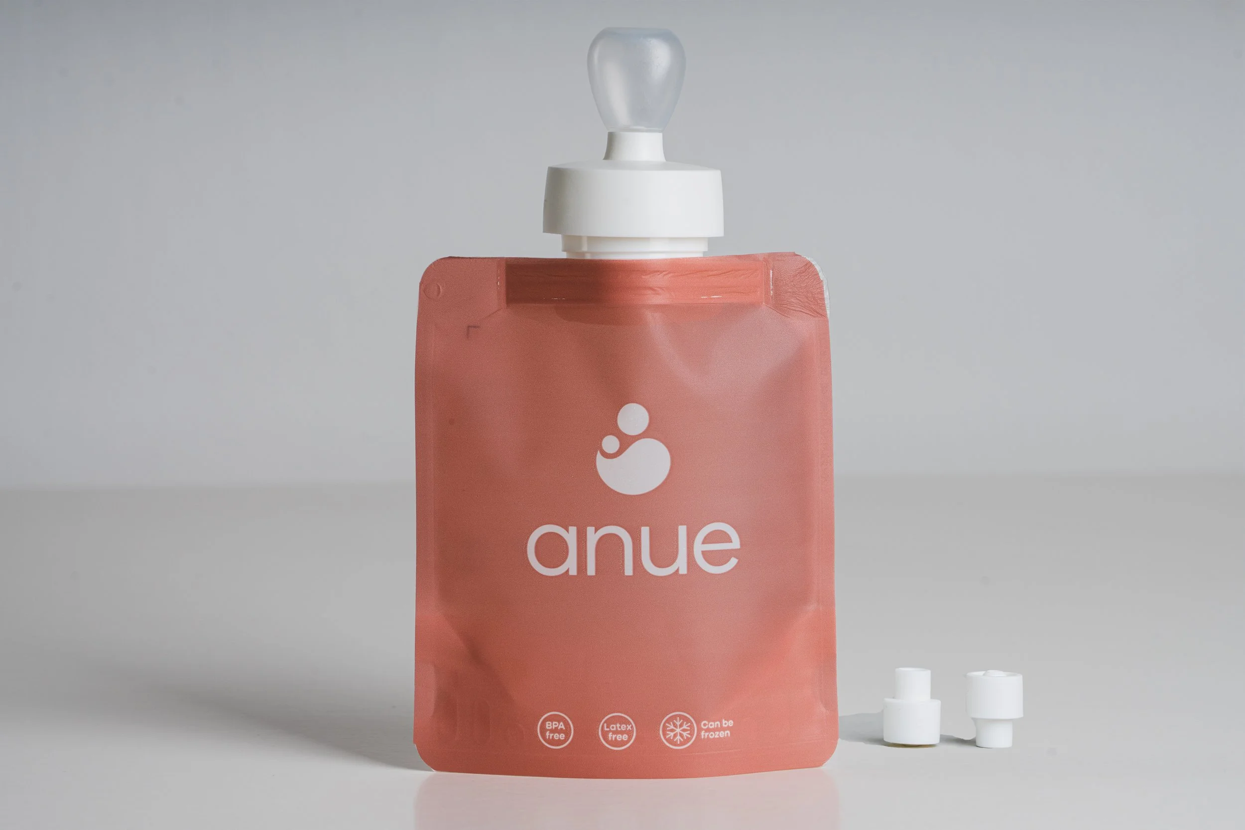 A pink flexible pouch with the brand name 'anue' and a logo, containing baby food, with a white screw cap and two additional white bottle collars beside it.
