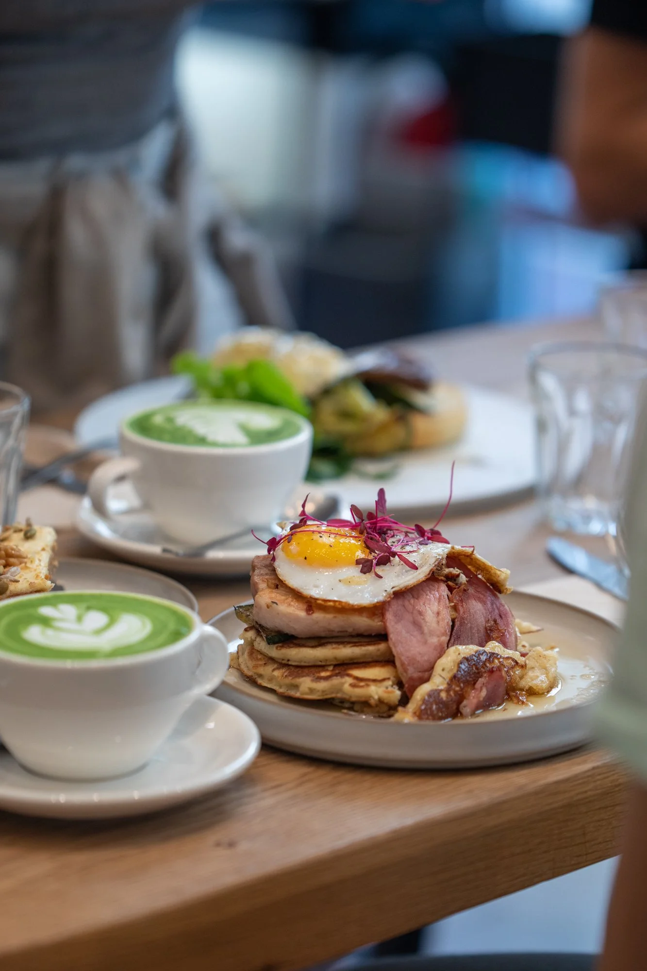 Close-up of breakfast dishes on a wooden table, featuring a stack of pancakes topped with bacon, a fried egg, and microgreens, accompanied by two cups of green matcha lattes.