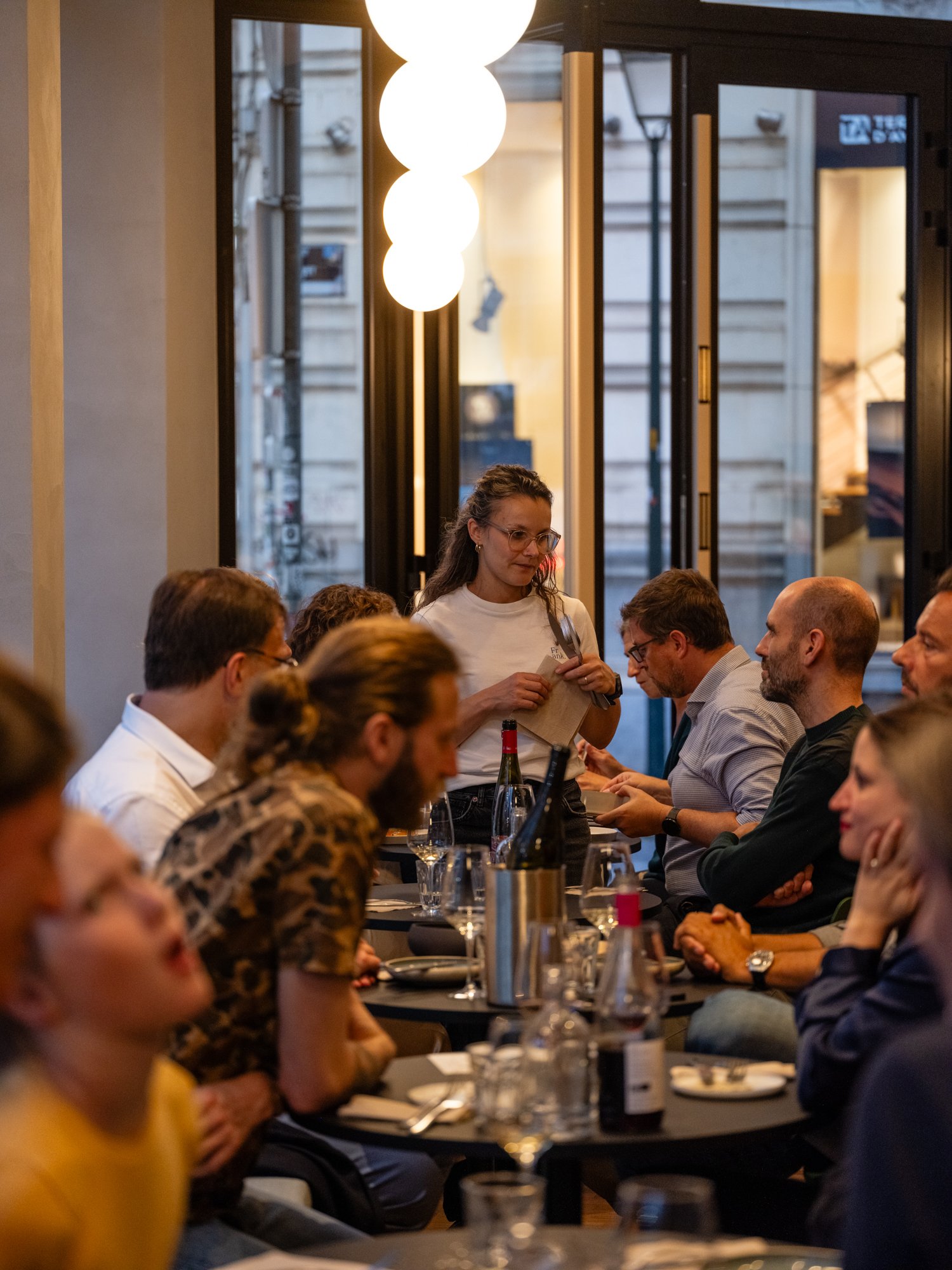 Group of people sitting and talking at a restaurant table indoors, with a server taking an order. Bottles of wine and glasses are on the tables, and large hanging lights are visible.