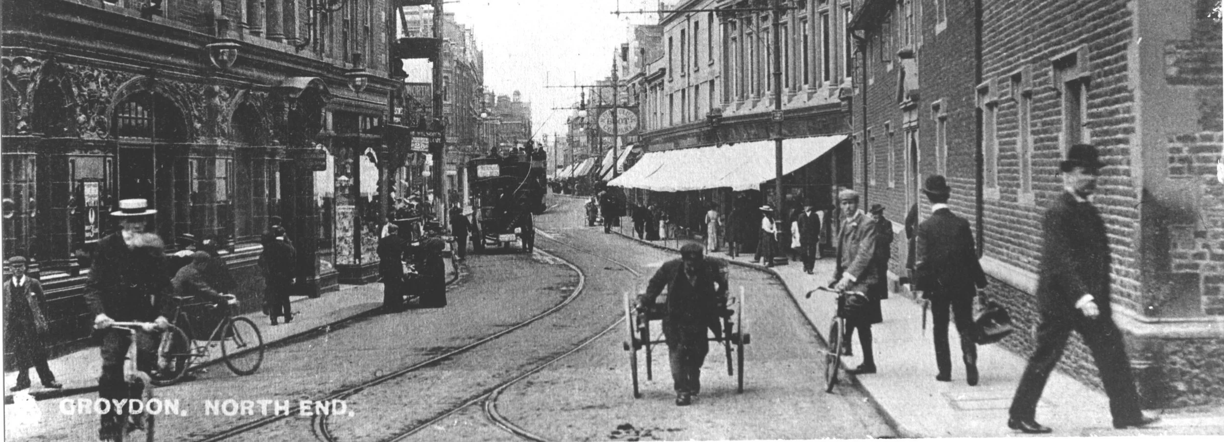 Black and white photo of Croydon's North End. Men in suits and hats are walking, cycling and one is pulling a cart. Women in Edwardian dress are browsing and walking past shops.