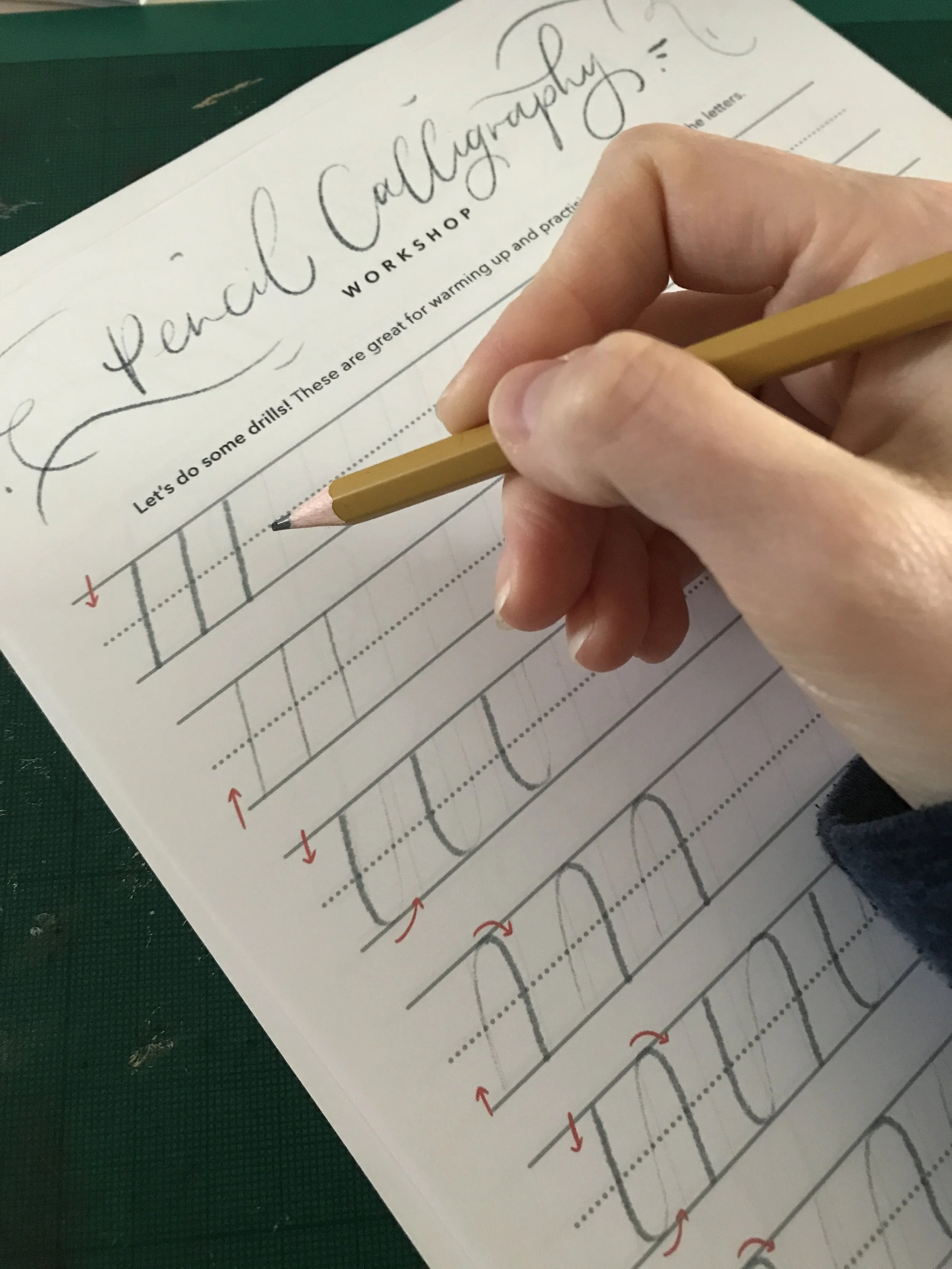 A person practicing handwriting on a worksheet titled 'Penal Calligraphy Workshop.' The worksheet has dotted guide lines and arrows indicating stroke directions. The person is holding a yellow pencil over the worksheet.