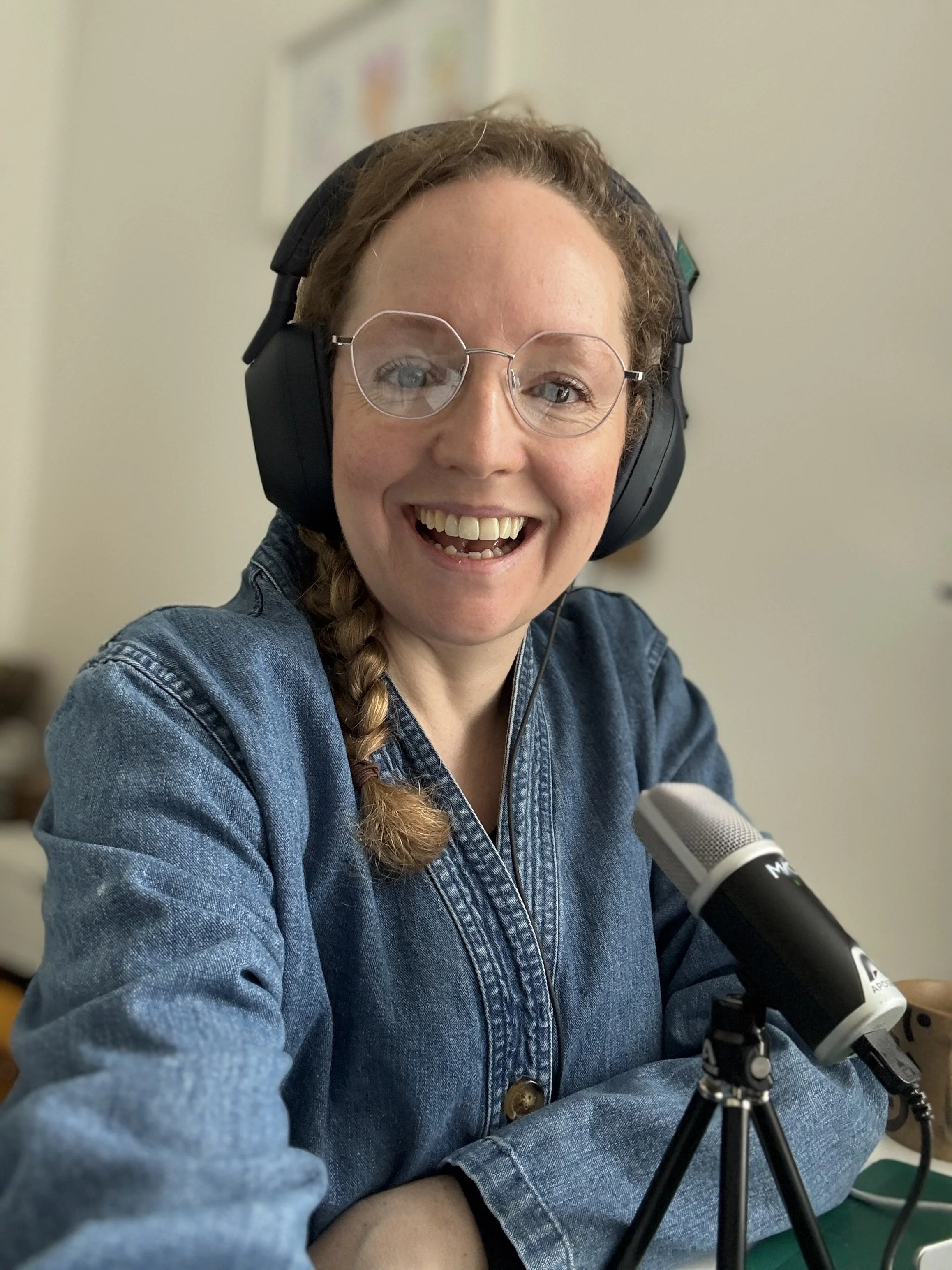 A woman with glasses, braided hair, and headphones smiling at a microphone.