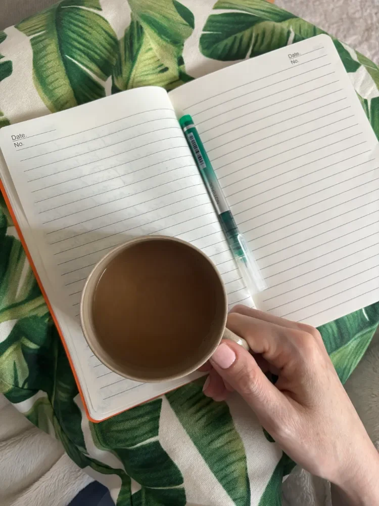 Laura's hand holding a mug of coffee with journal notebook open and pen ready