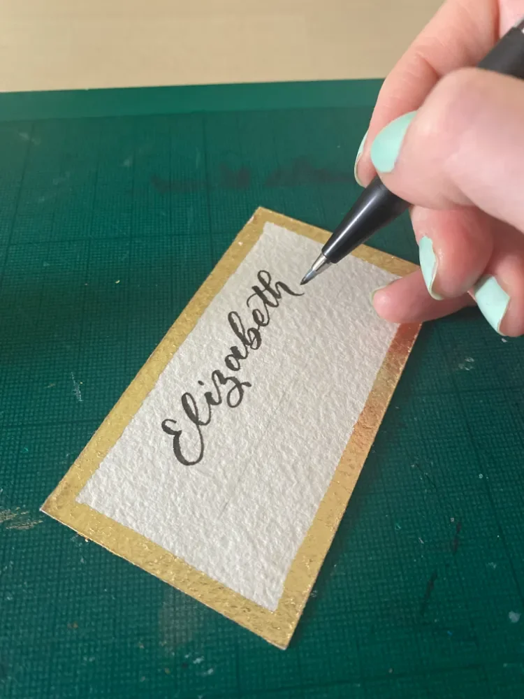 Elizabeth being written in brush script calligraphy on a gold-edged name card