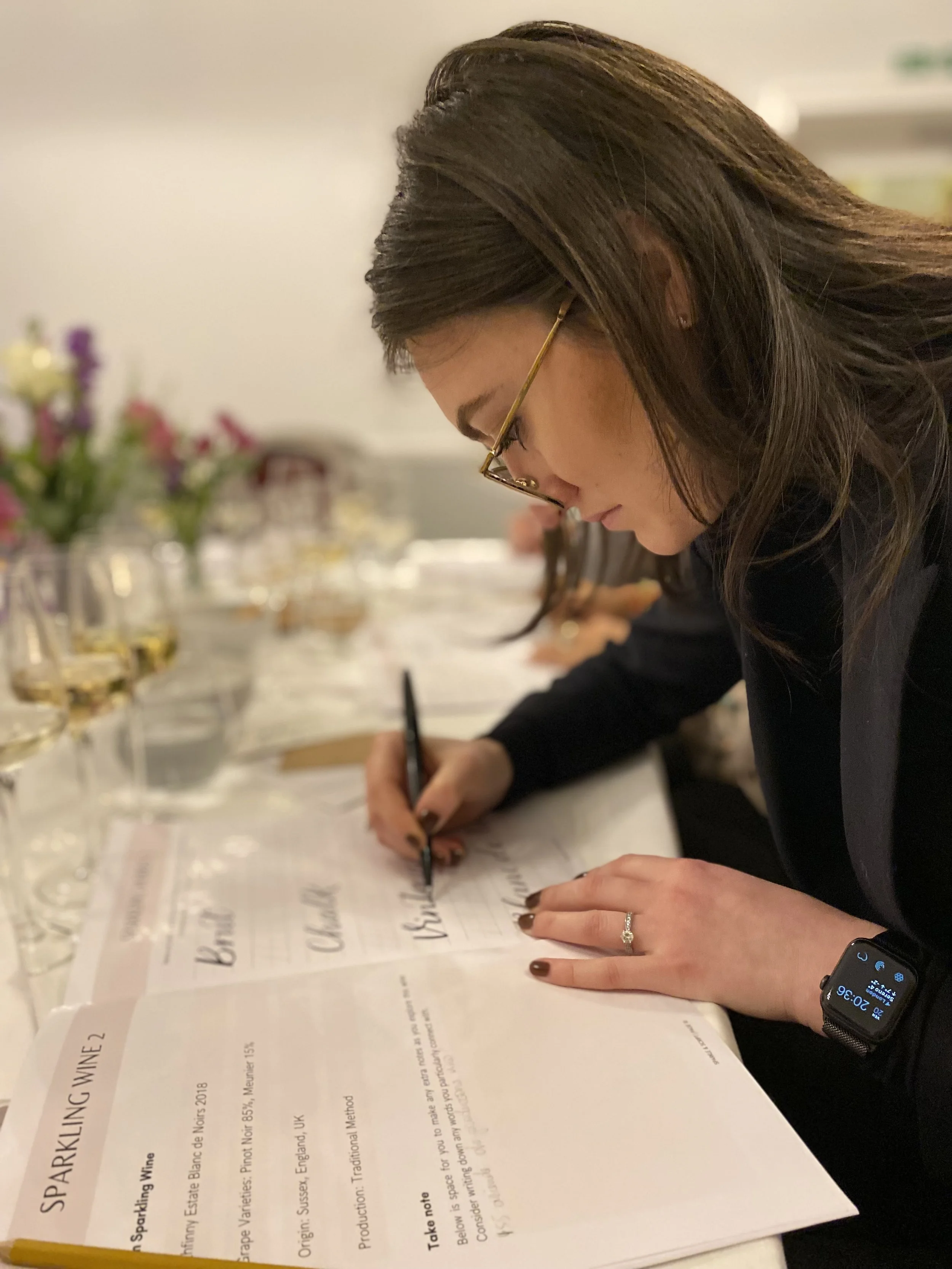 A woman wearing glasses and a smartwatch writes on a tasting sheet at a wine tasting event with wine glasses and flowers in the background.