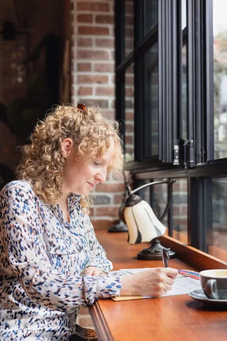A woman with curly blonde hair sitting at a wooden bench seat, practicing calligraphy, beside a window, a lamp, and a coffee cup.