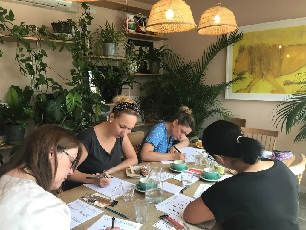 Four women sitting at a wooden table practicing calligraphy in a calligraphy workshop, at The Printworks London in a cozy, plant-filled room.
