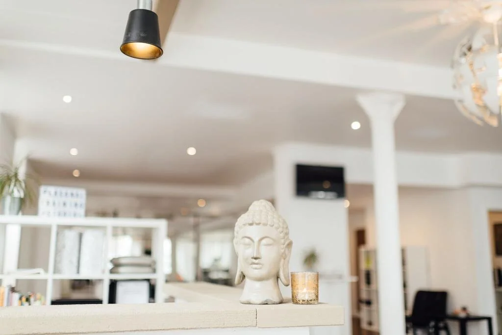 Innenaufnahme eines modernen, hellen Wohnzimmers mit weißen Wänden und Möbeln, einer schwarzen Pendelleuchte, einer weißen Säule und einer Buddha-Statue auf einem Regal.
