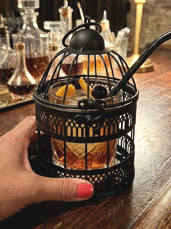 Black mini bird cage with cut glass cocktail glass inside with hand holding the cage